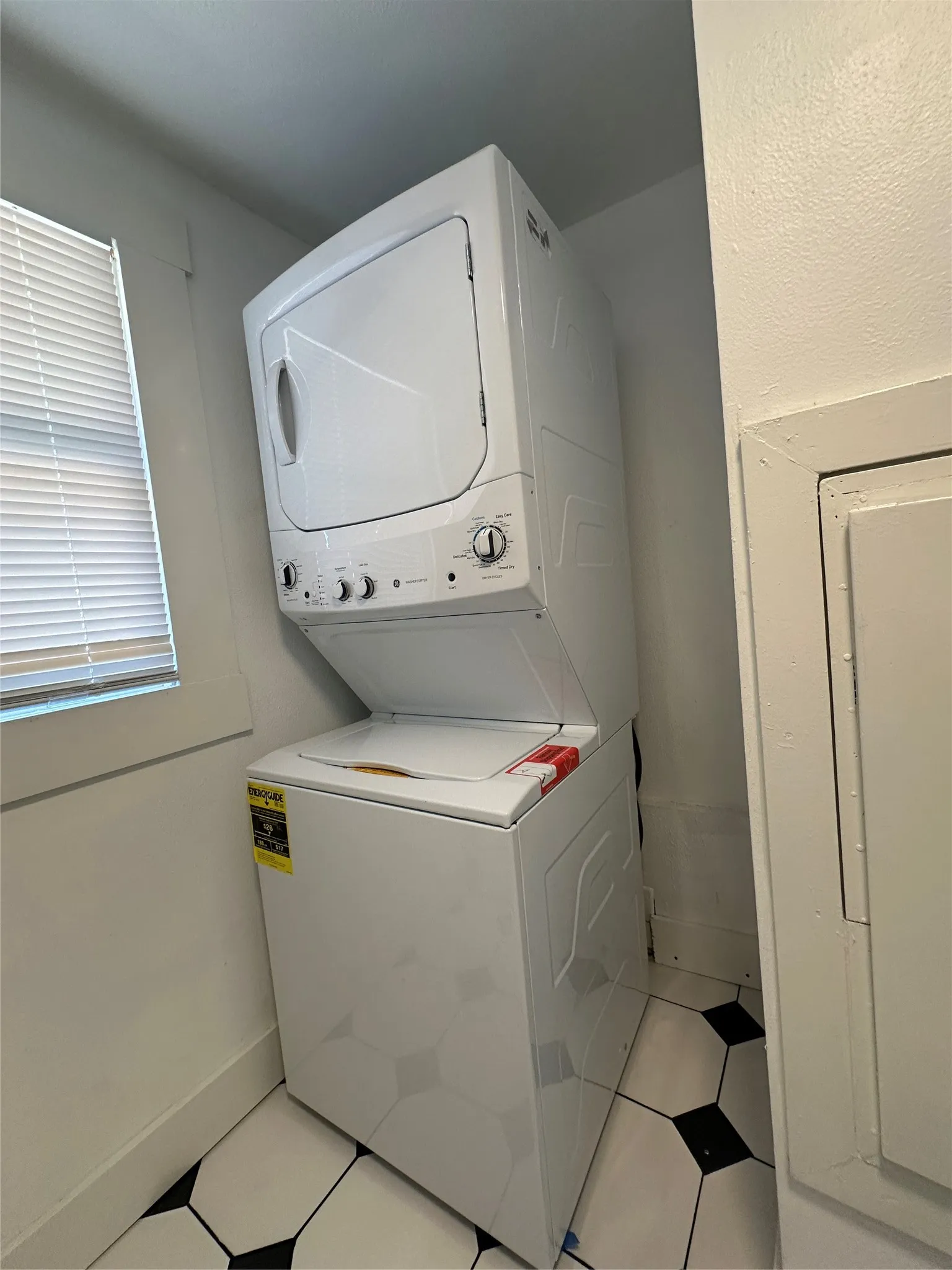 Stackable white washer and dryer unit, window with blinds, and geometric tile flooring