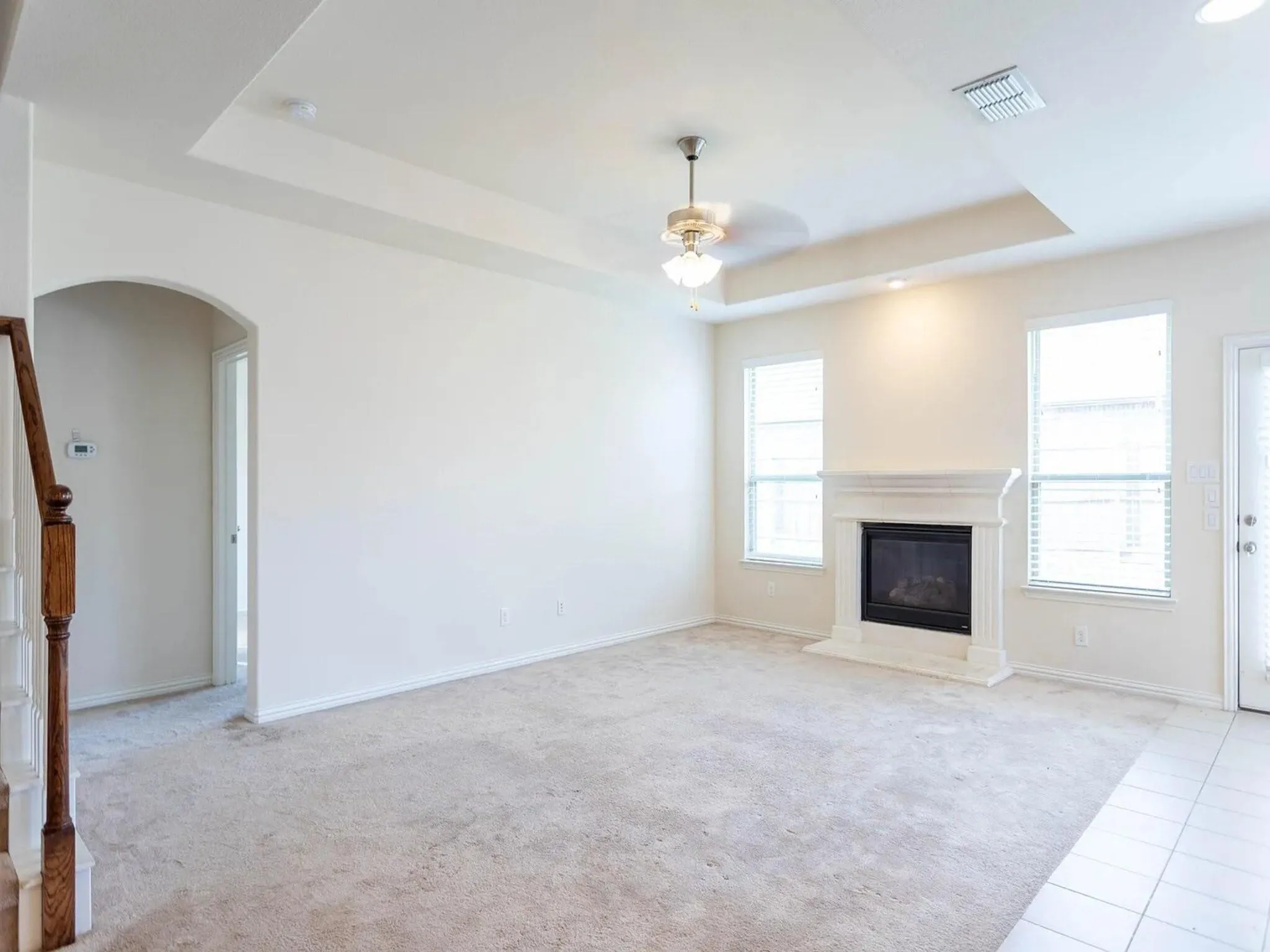 Recessed ceiling with a central fan light, a fireplace with a stone-finish mantle, multiple windows with blinds, and light-colored carpeting
