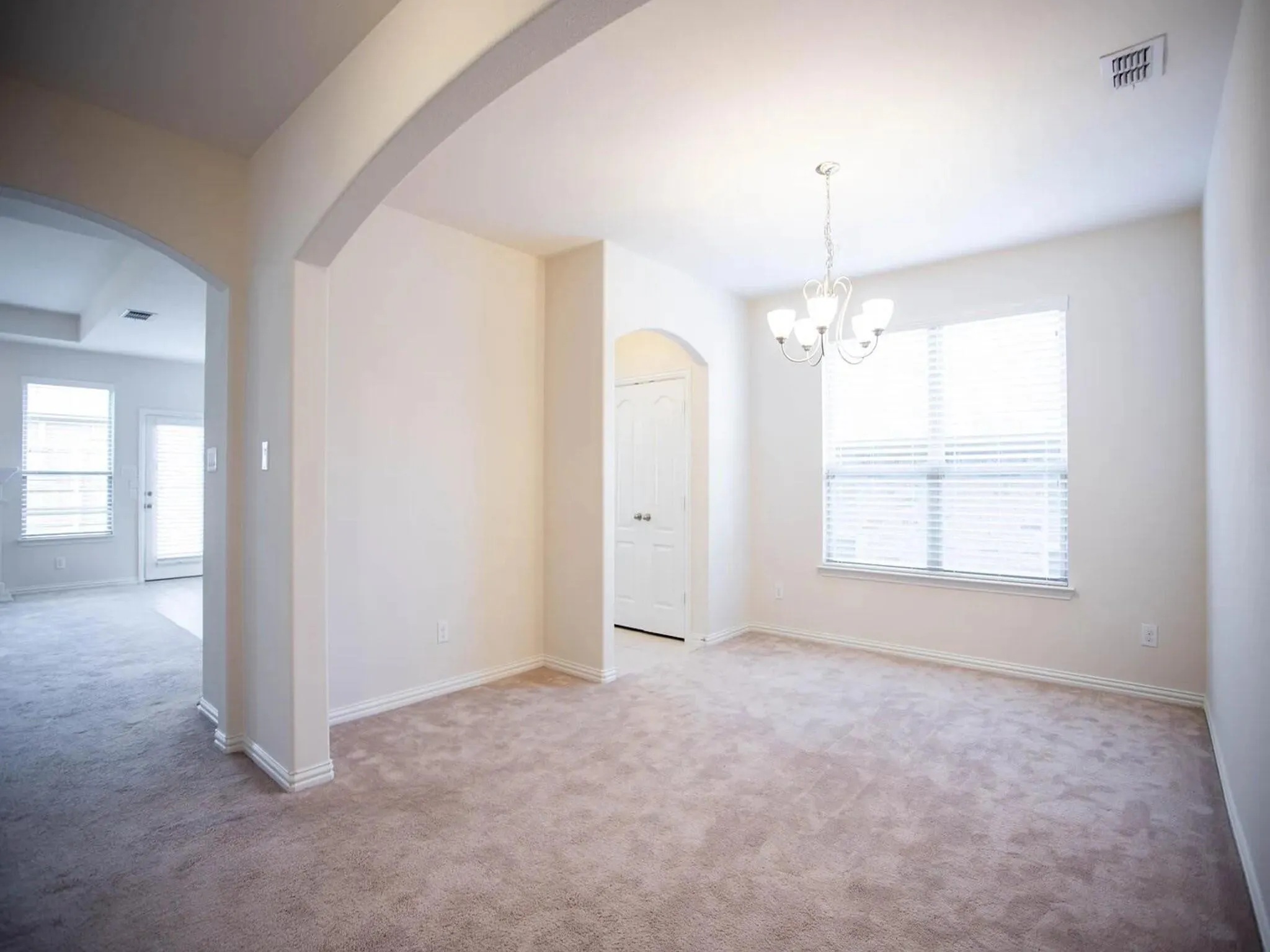 Carpeted interior space with arched entryway, overhead chandelier, and a large window with blinds
