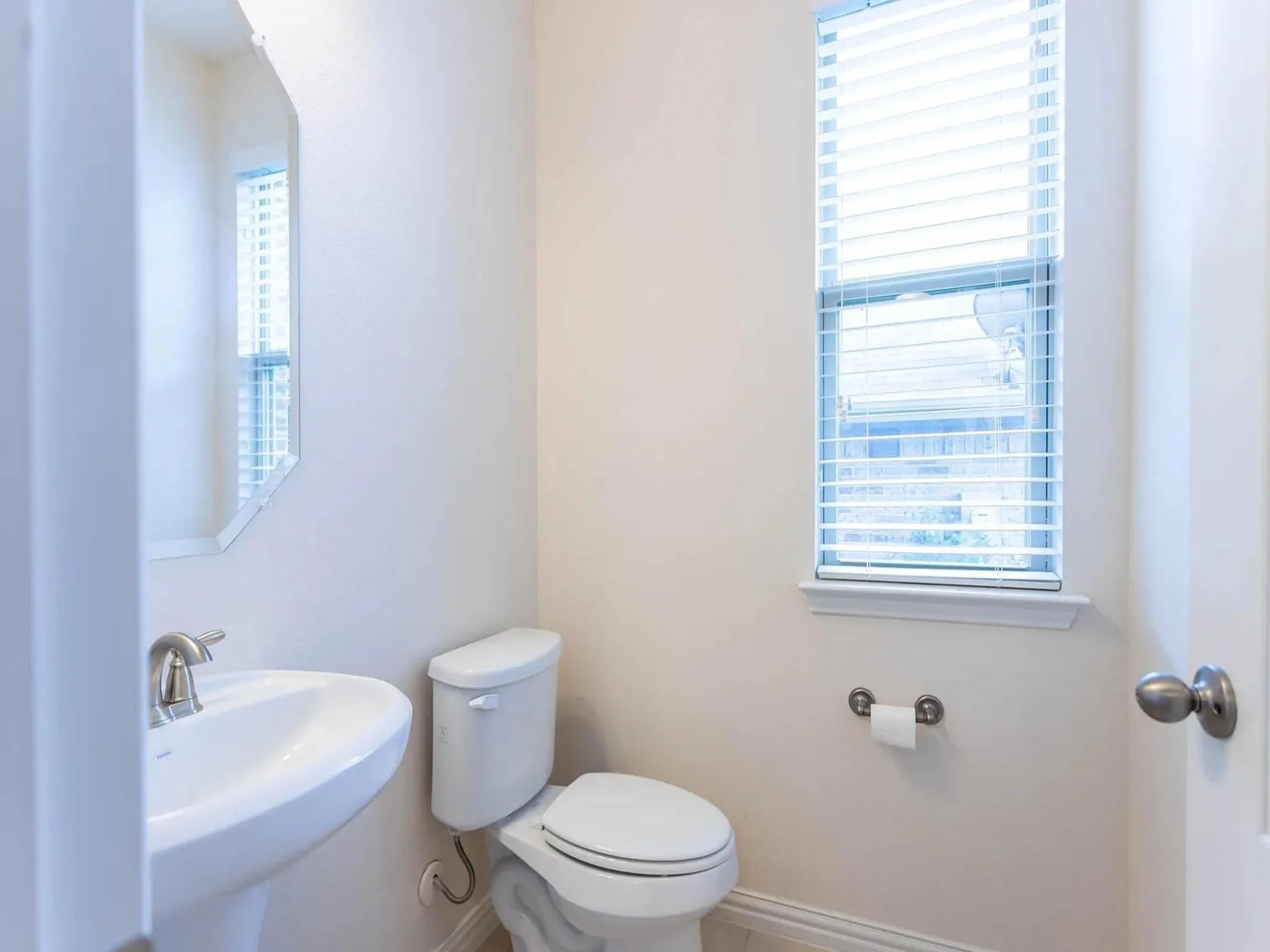 Pedestal sink with brushed nickel faucet, wall-mounted mirror, toilet, and window with blinds