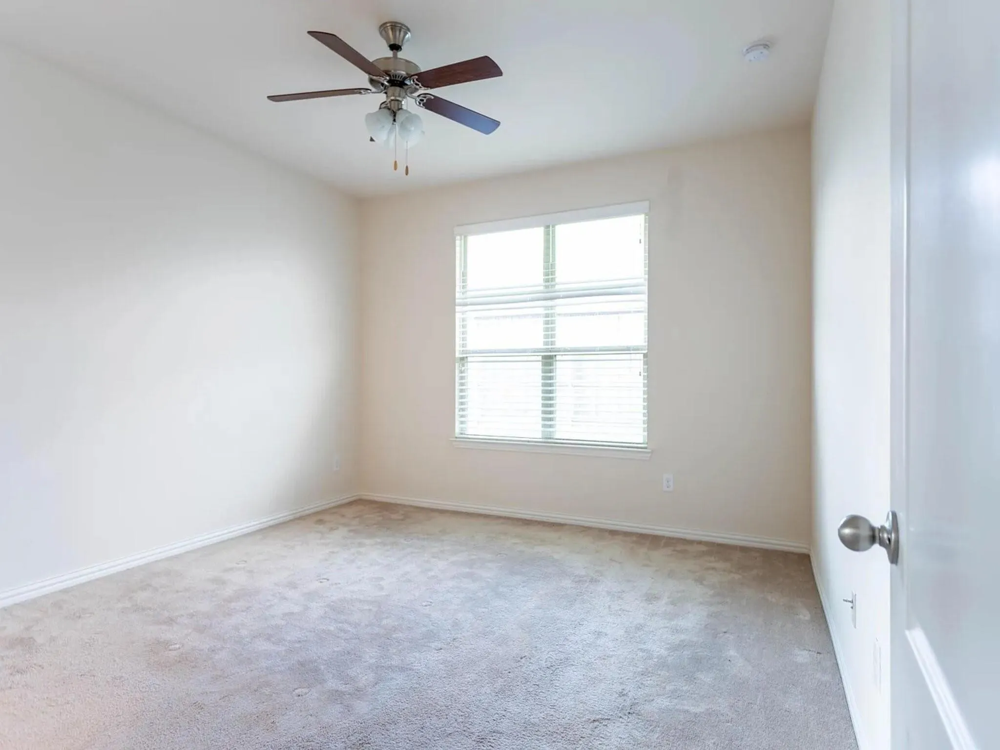 Neutral tone wall color, carpeted flooring, window with horizontal blinds, and a ceiling fan with integrated lighting