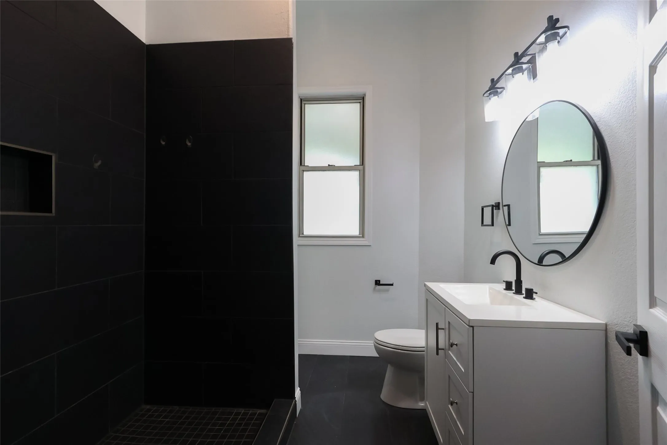 Bathroom featuring a white vanity with a white countertop, a black-framed circular mirror, a black tiled shower with an integrated niche, a white toilet, and black-finish hardware