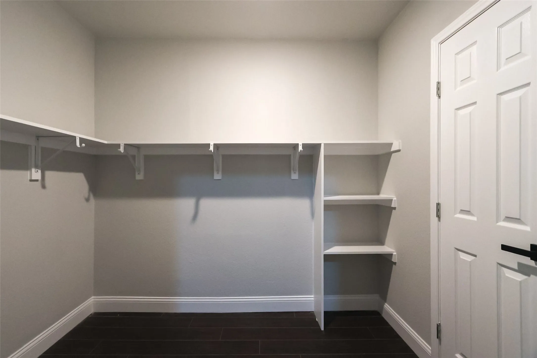 Spacious walk-in closet featuring white built-in shelving, dark wood-finish flooring, light grey wall paint, and a white paneled door with matte black hardware
