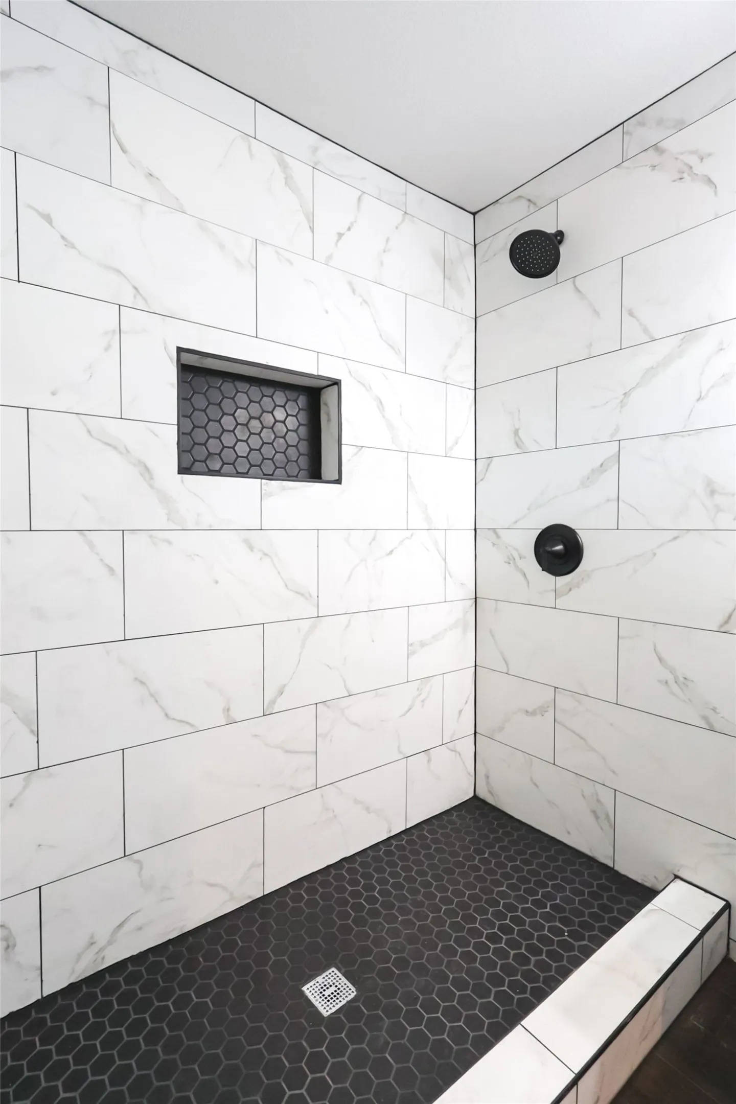 Shower with white stone-finish subway tile walls, black hexagonal floor tile, matte black shower fixtures, and a built-in tiled niche