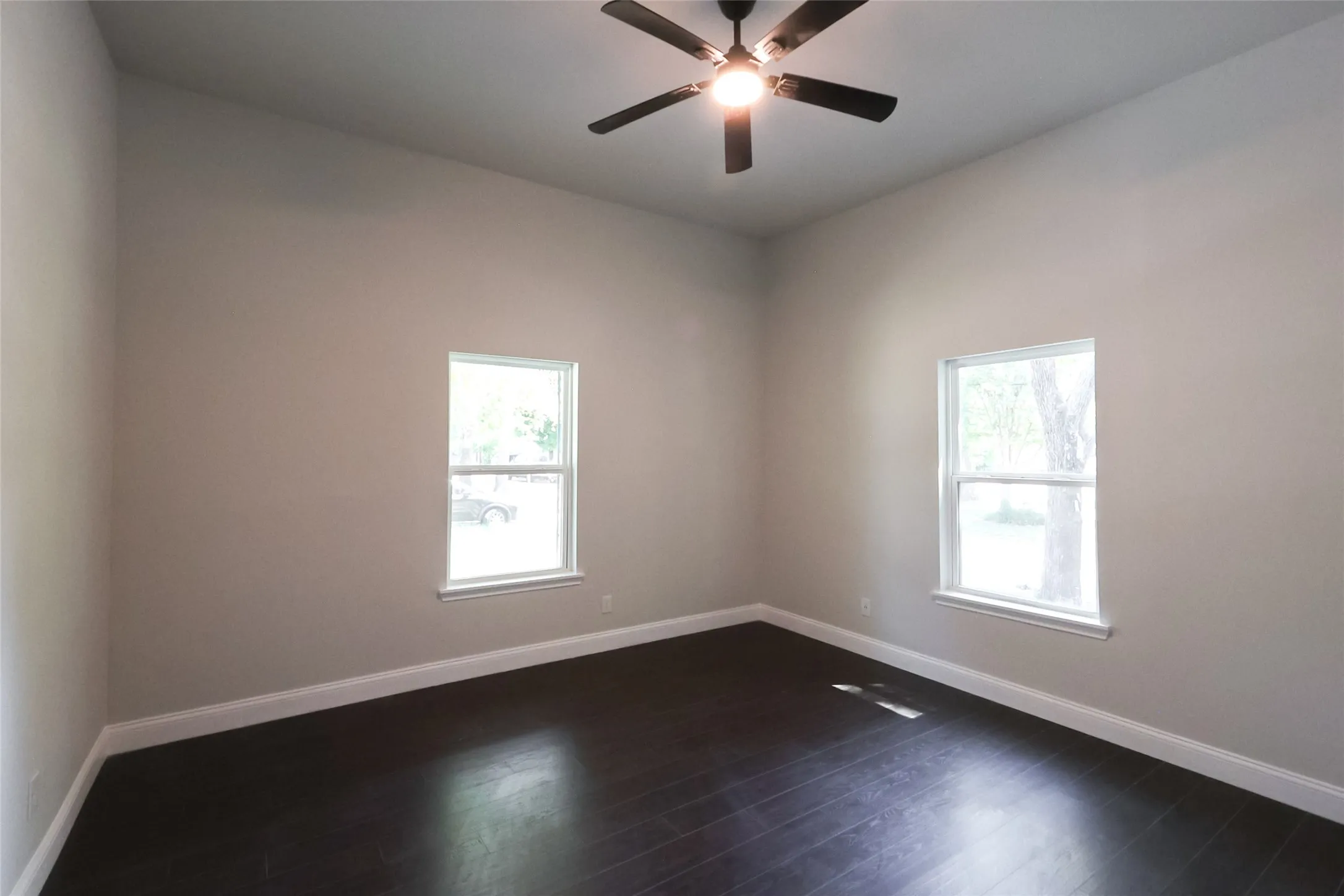 Dark wood-finish flooring, light neutral wall tones, white trim, two single-hung windows, and a ceiling fan with integrated lighting