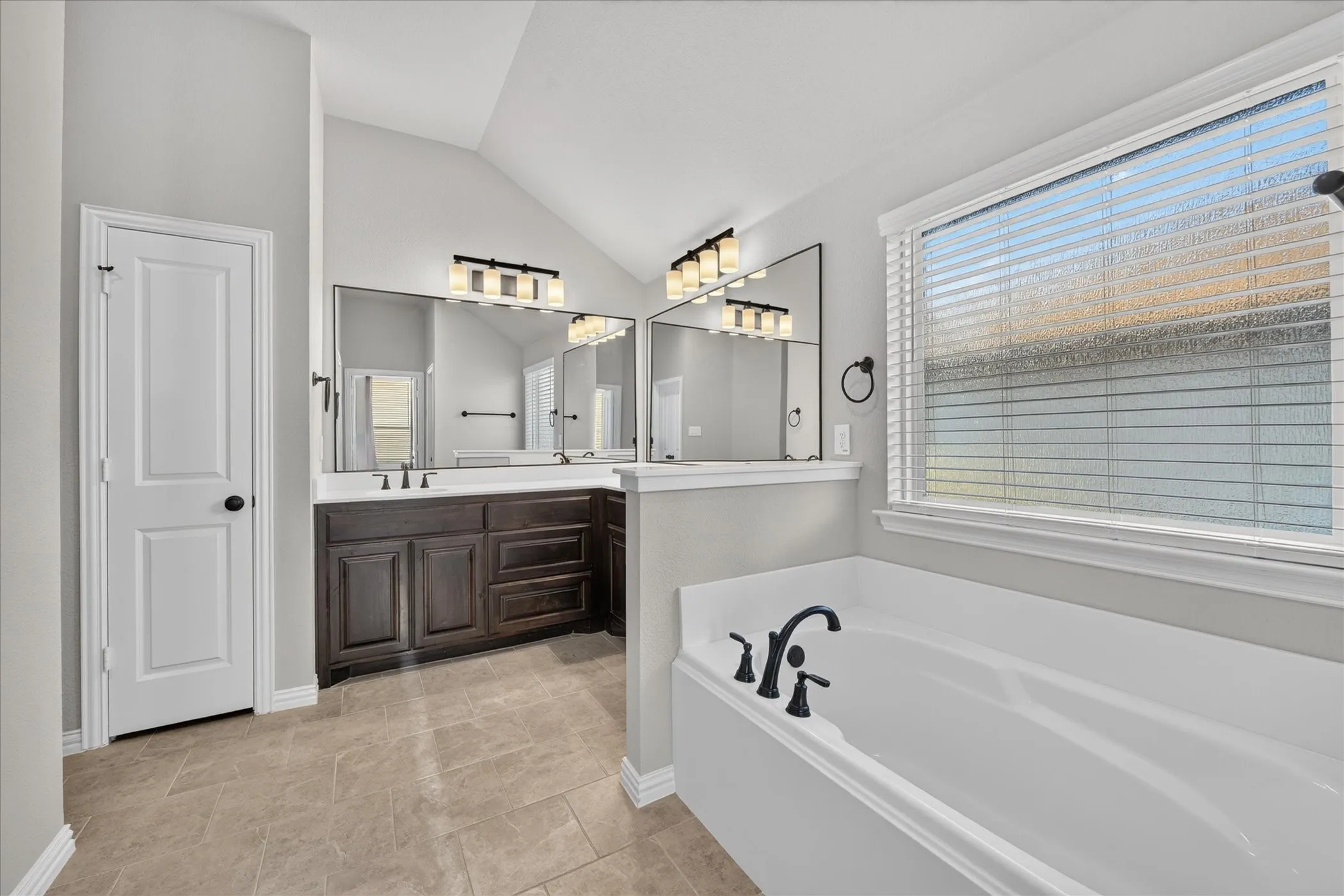 Vaulted ceiling, dual vanity with dark wood cabinetry, white countertop, large soaking tub with matte black fixtures, and tiled flooring