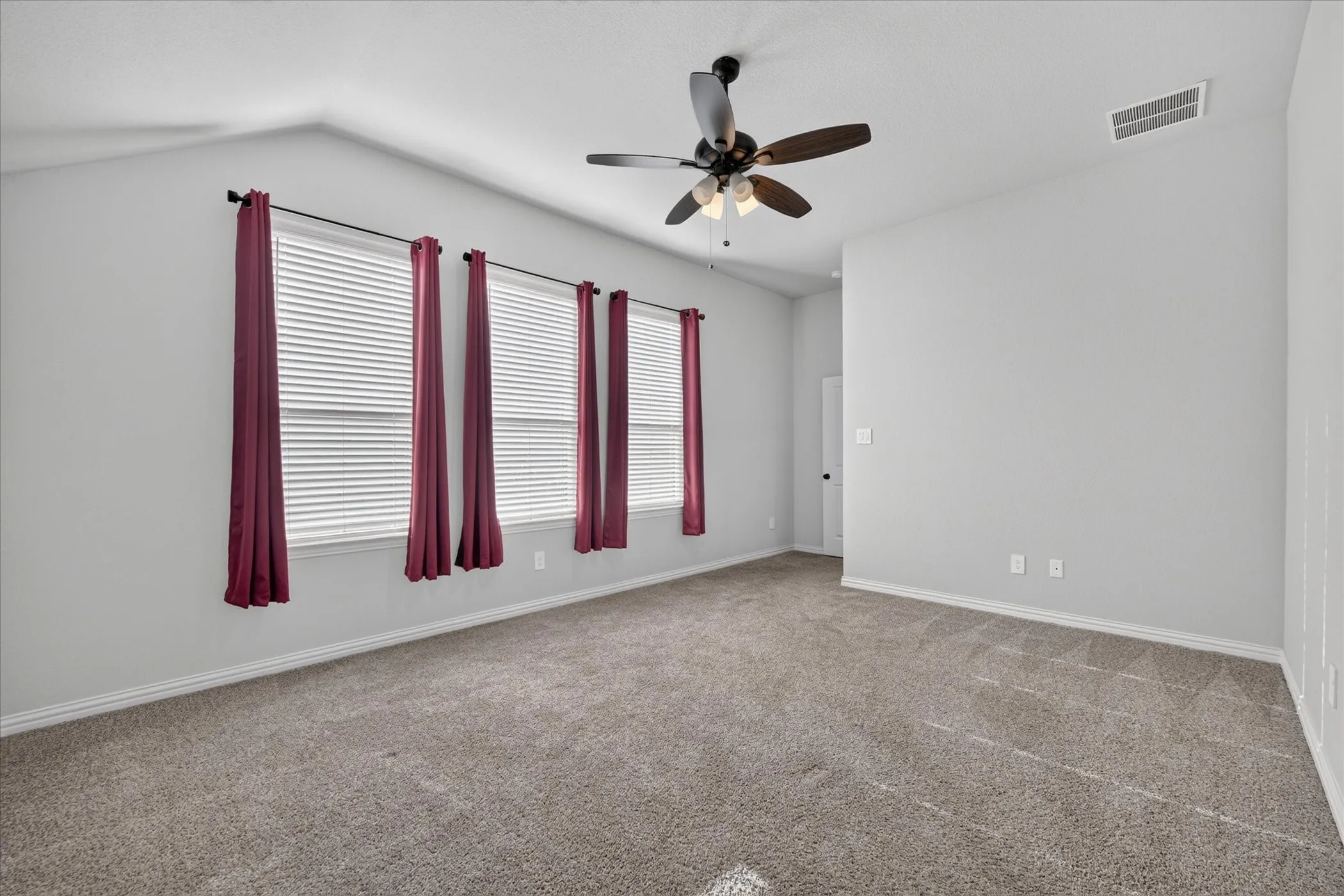Vaulted ceiling, ceiling fan with light fixture, gray carpeting, multiple windows with blinds, and light gray wall paint