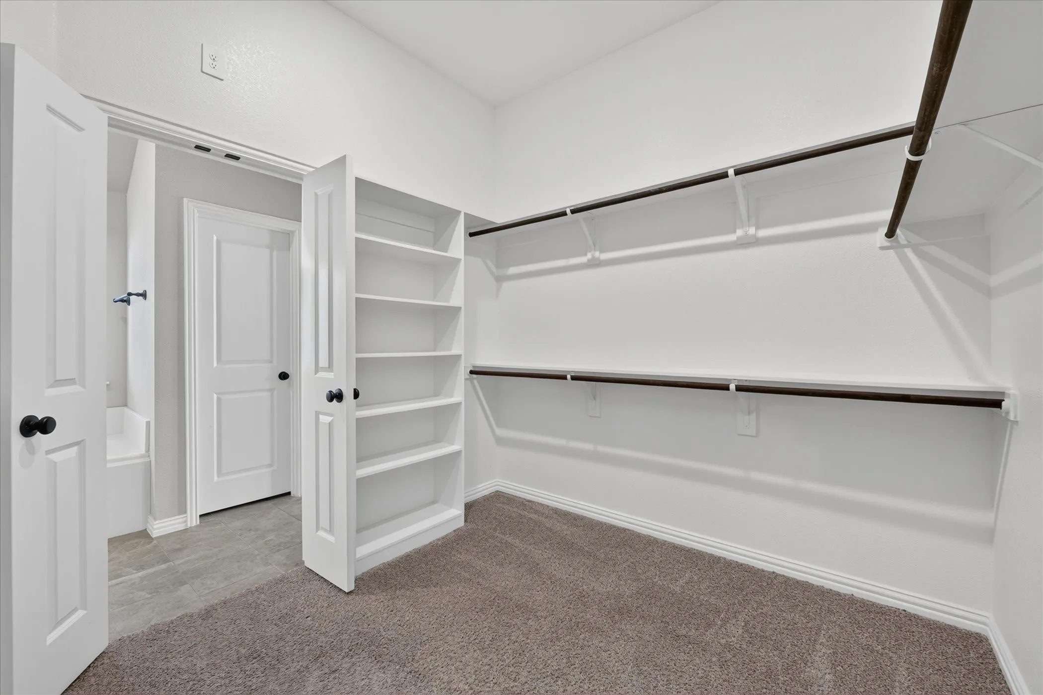 Carpeted flooring, built-in shelving, two rows of garment rods, white paneled doors, and matte black hardware