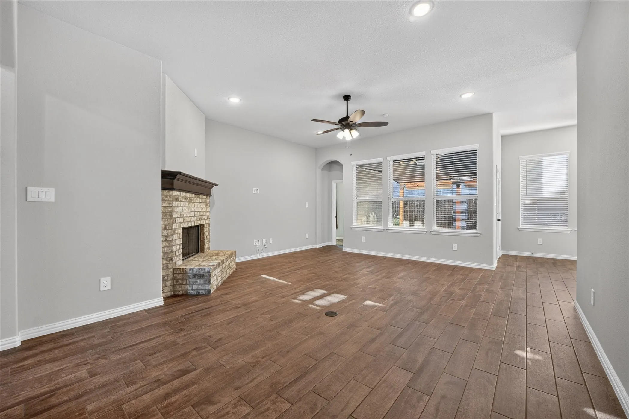 Spacious living area featuring wood-finish flooring, a brick fireplace with a dark wood mantel, multiple windows, and a ceiling fan