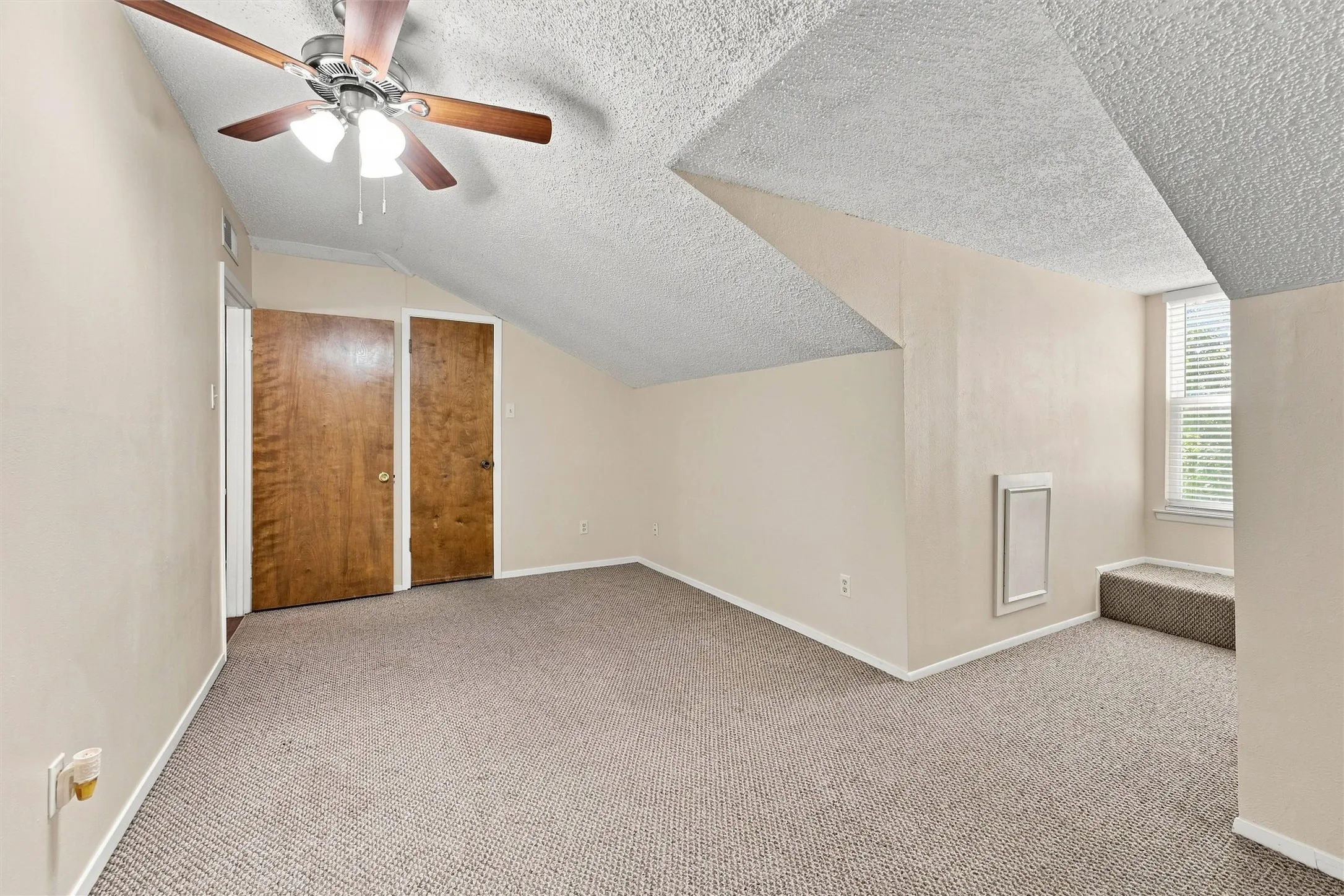 Sloped ceilings with textured finish, ceiling fan with light fixture, carpeted flooring, two wood-paneled doors, and a window with blinds