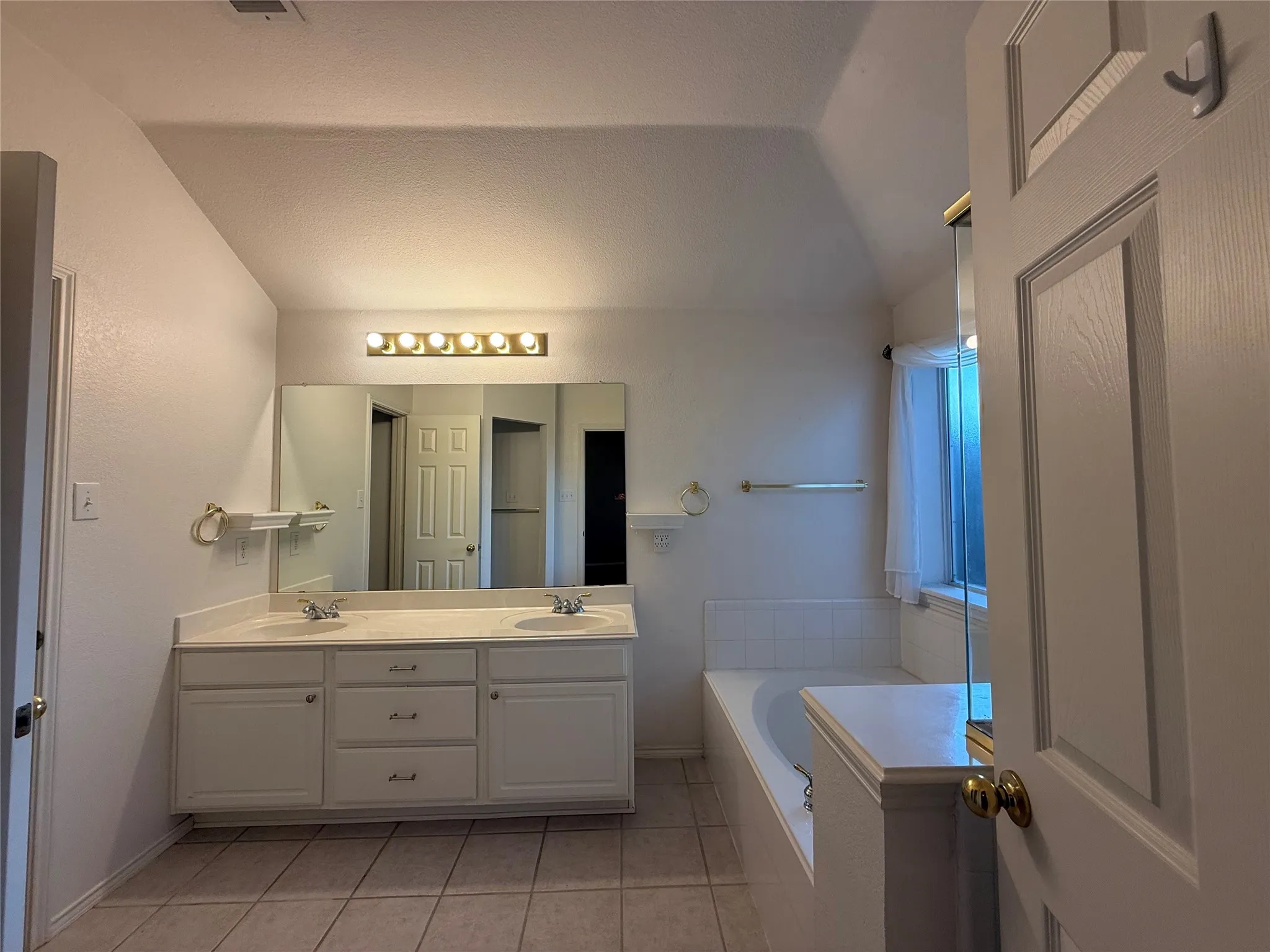 Dual vanity with integrated sinks, expansive wall-mounted mirror, overhead vanity lighting, tiled flooring, and a built-in bathtub with tiled surround