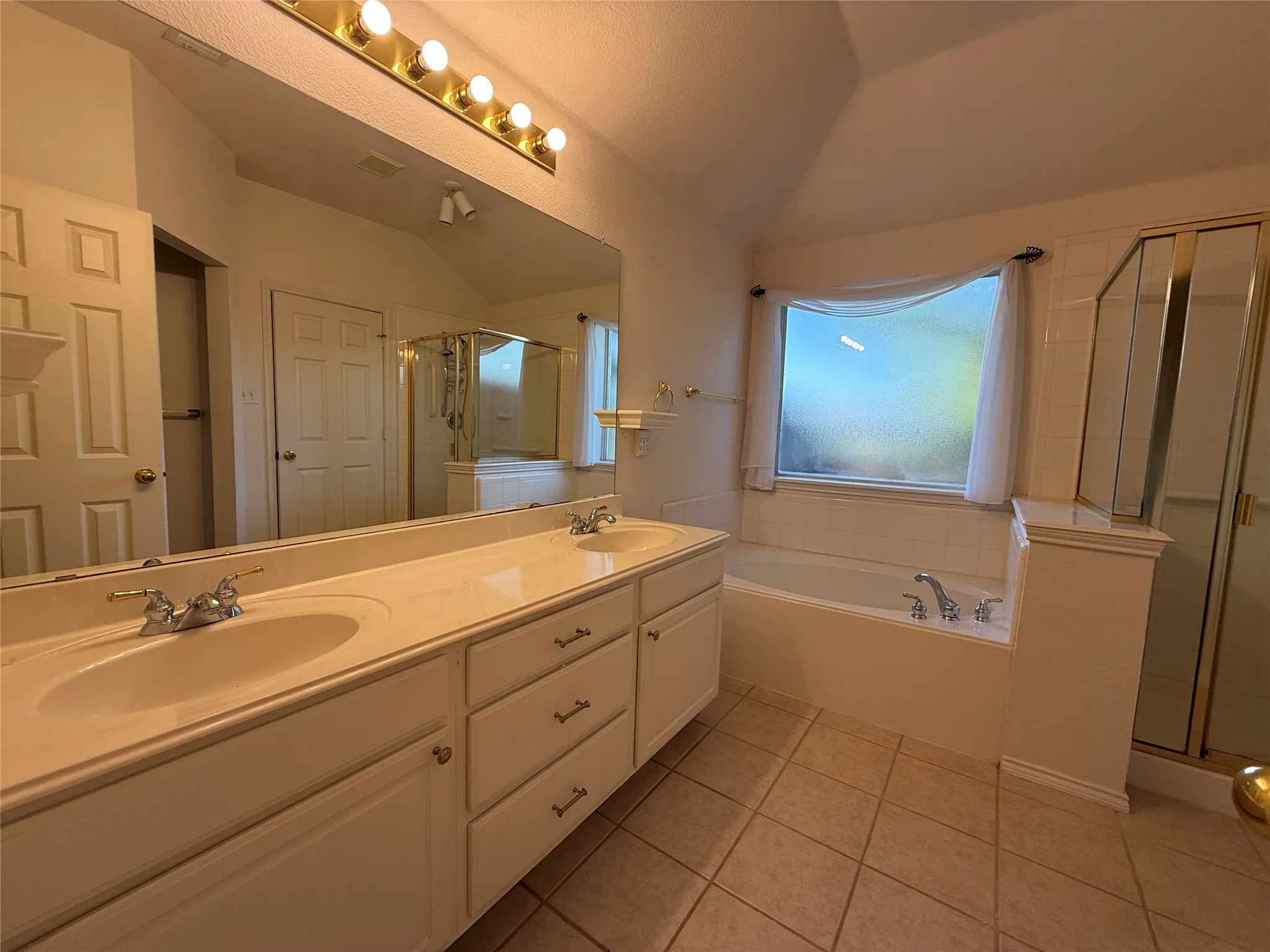 Dual vanity with integrated sinks, wide mirror, built-in soaking tub, separate glass-enclosed shower, and tile flooring