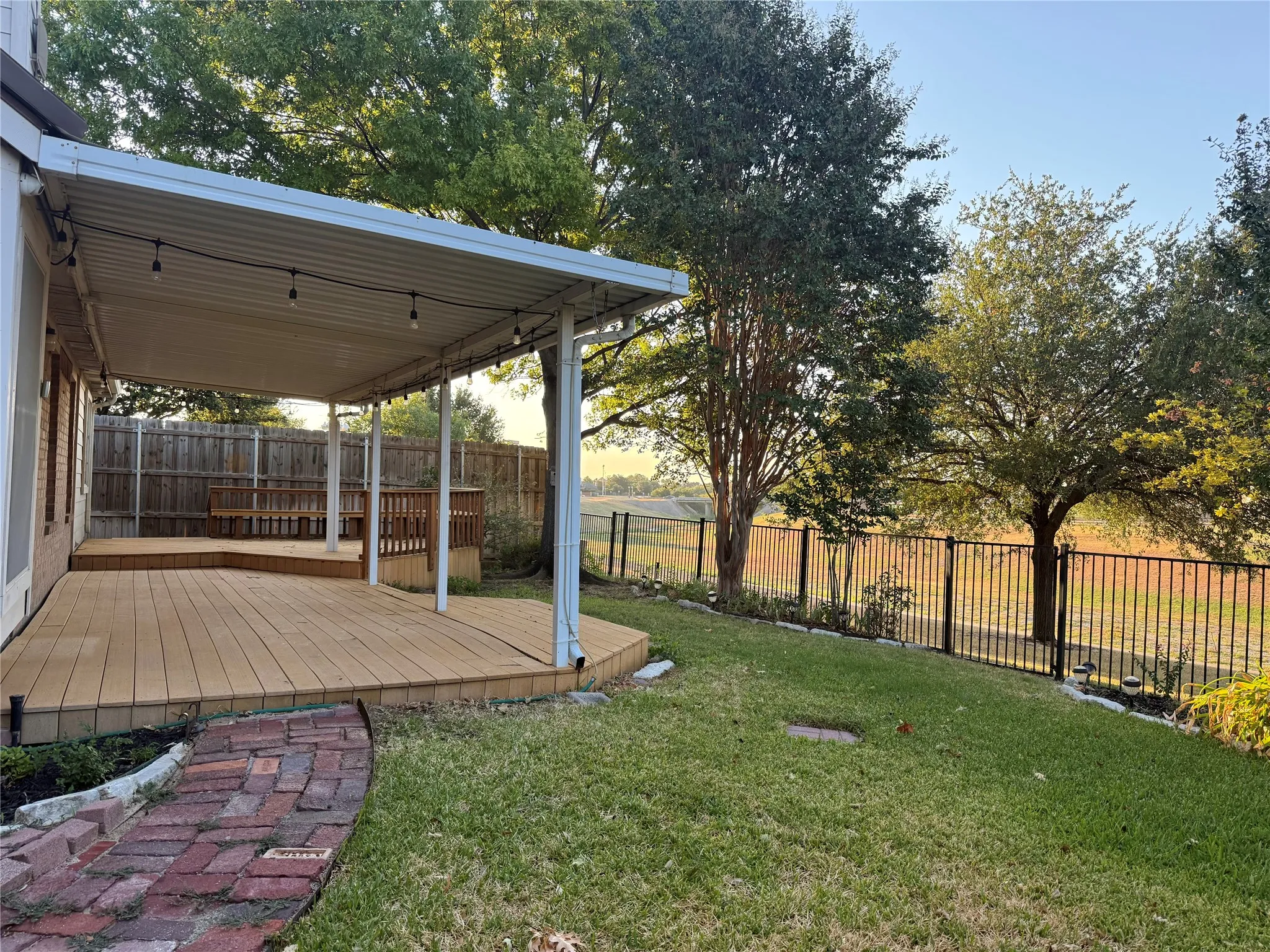 Expansive backyard featuring a multi-level wood-finish deck, covered patio, mature trees, and wrought iron fencing