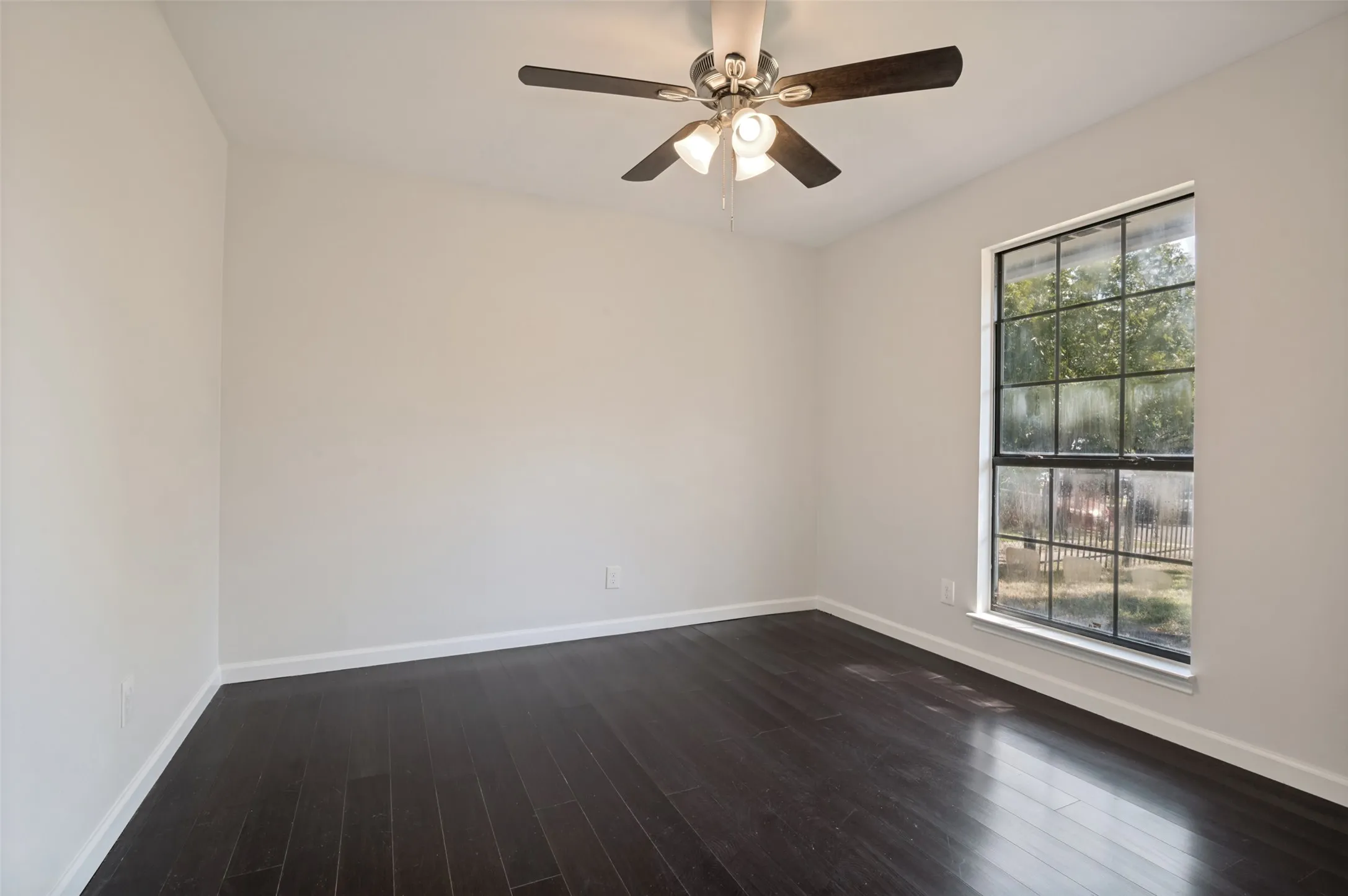 Spacious room featuring dark wood-finish flooring, a large multi-pane window, and a ceiling fan with integrated lighting