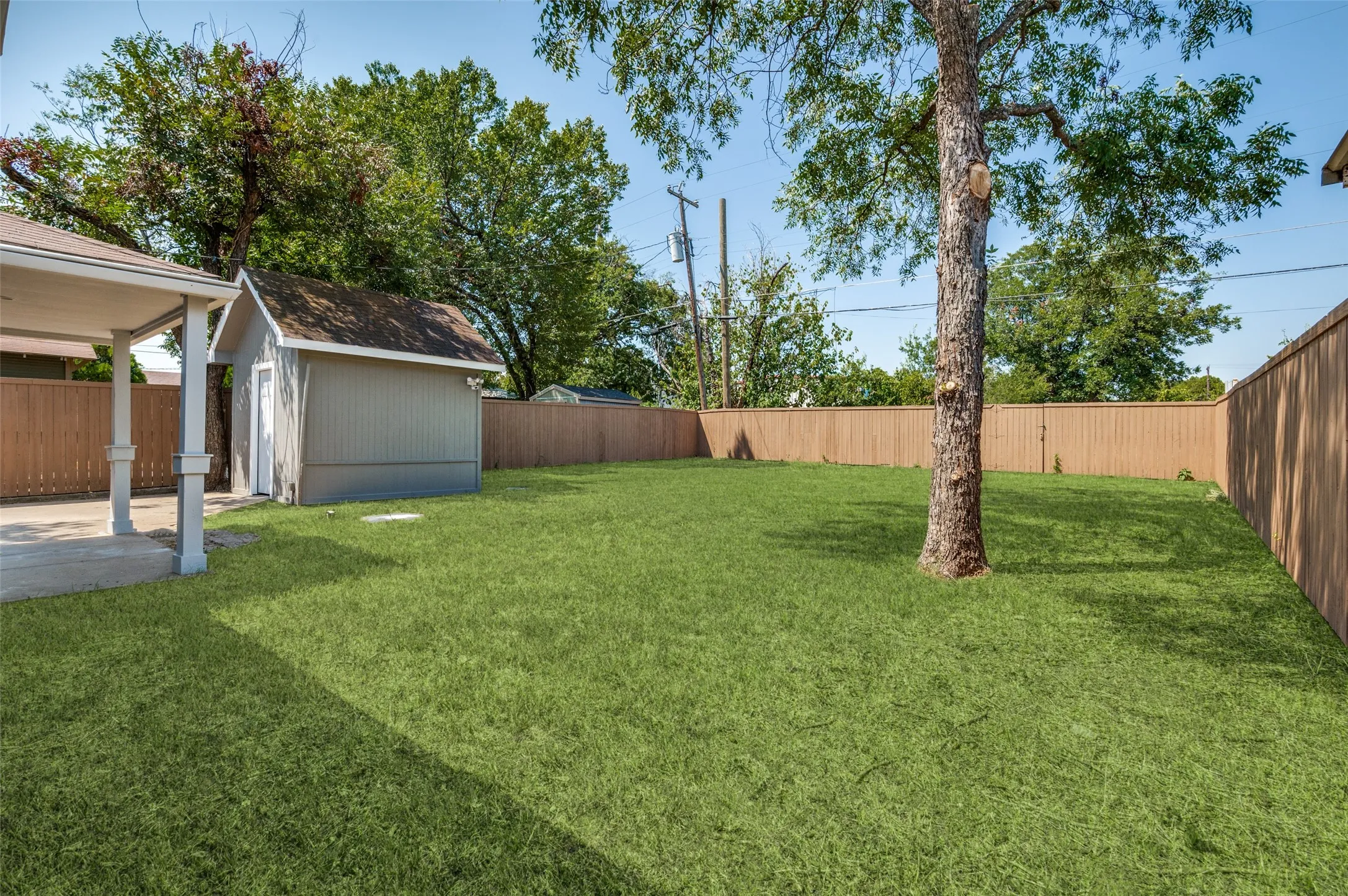 Spacious backyard featuring a manicured lawn, established tree, wood privacy fencing, a covered patio, and a storage shed