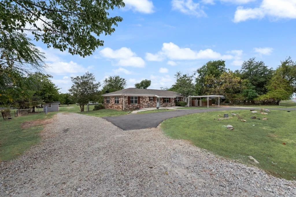 Brick exterior residence with a covered carport, expansive gravel driveway, and a separate paved parking area