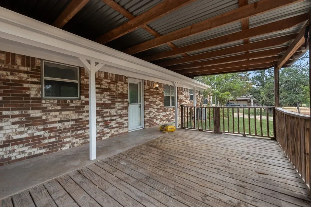 Expansive covered porch featuring a wooden deck, rustic wooden railing, exposed wood beam ceiling, and corrugated metal roof