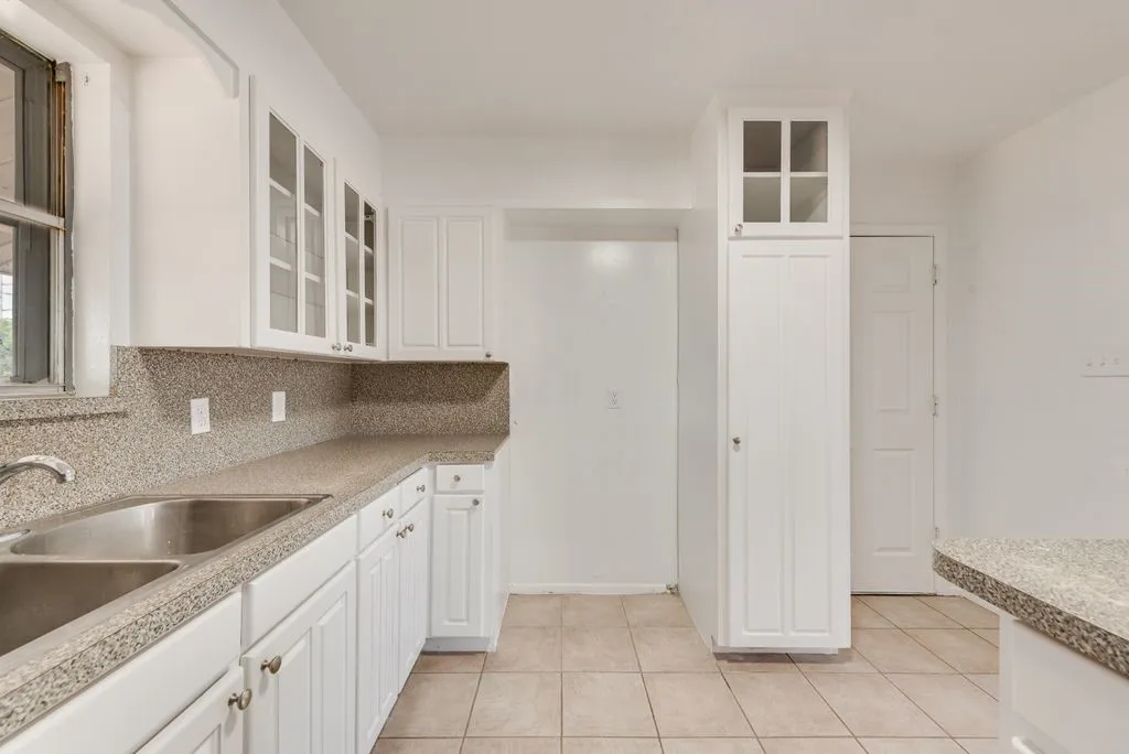 White shaker-style cabinetry with glass-front upper cabinets, speckled countertops with integrated backsplash, and a double basin stainless steel sink