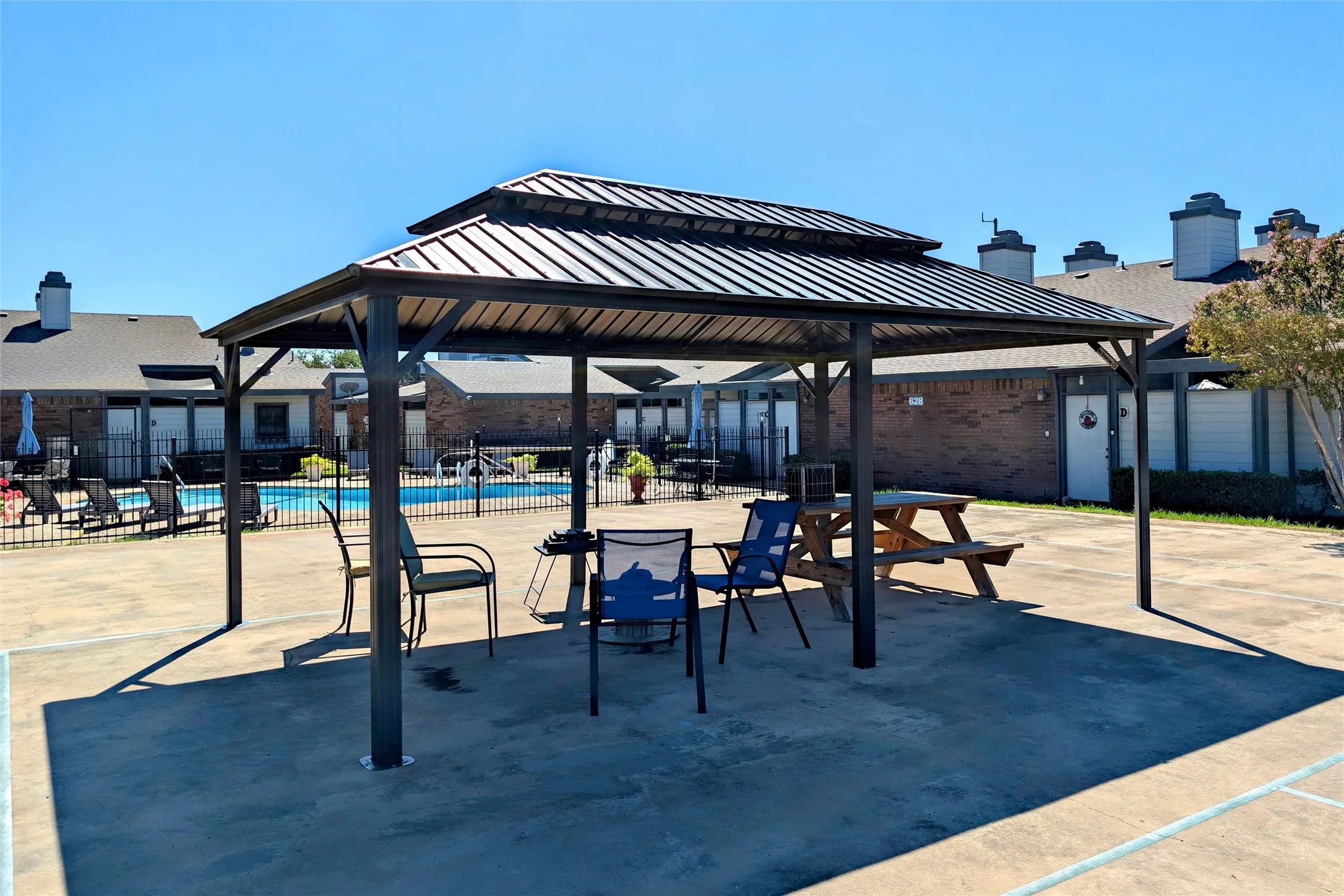 Outdoor gazebo has tiered metal roof, concrete patio, and a Community swimming pool beyond a black metal fence