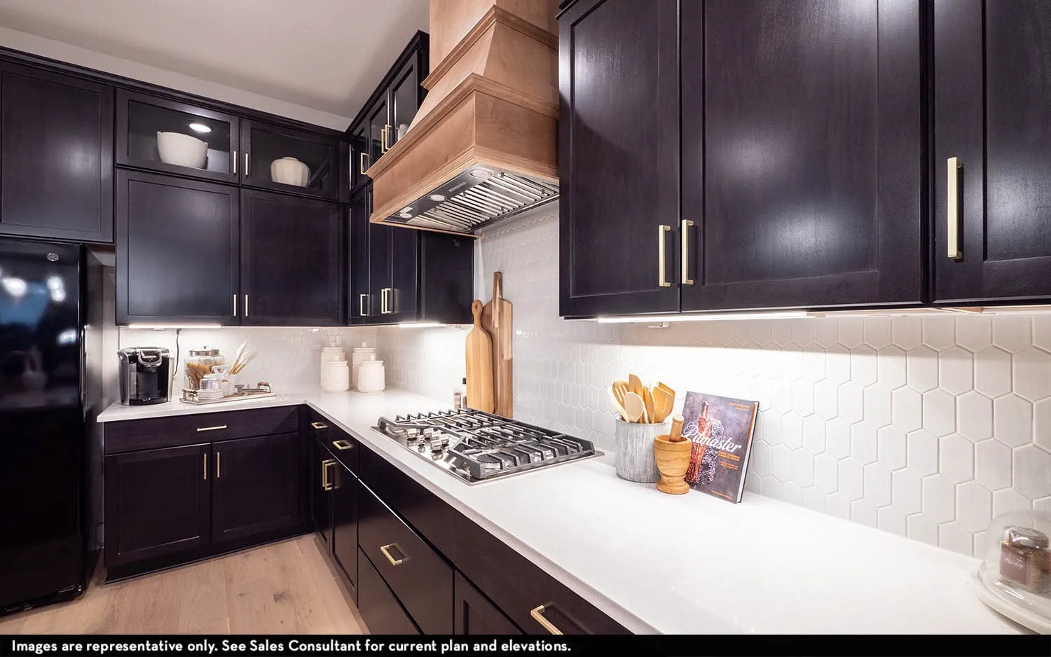 Black cabinetry with gold-finish hardware, stainless steel gas cooktop, light wood-finish range hood, white countertop, and geometric tile backsplash