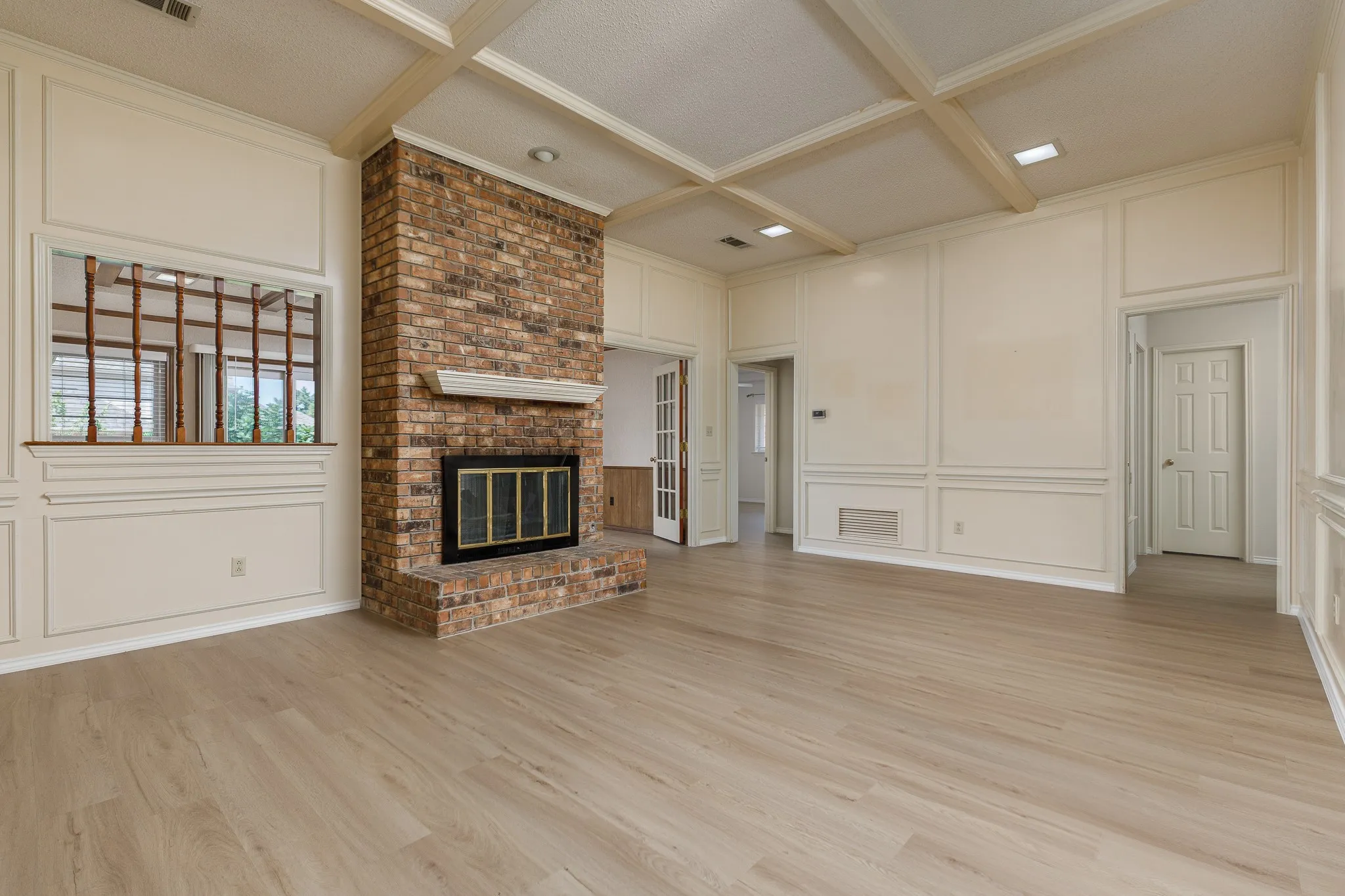 Expansive room featuring a brick fireplace, wood-finish flooring, coffered ceiling, wall paneling, and an interior window with wood detailing