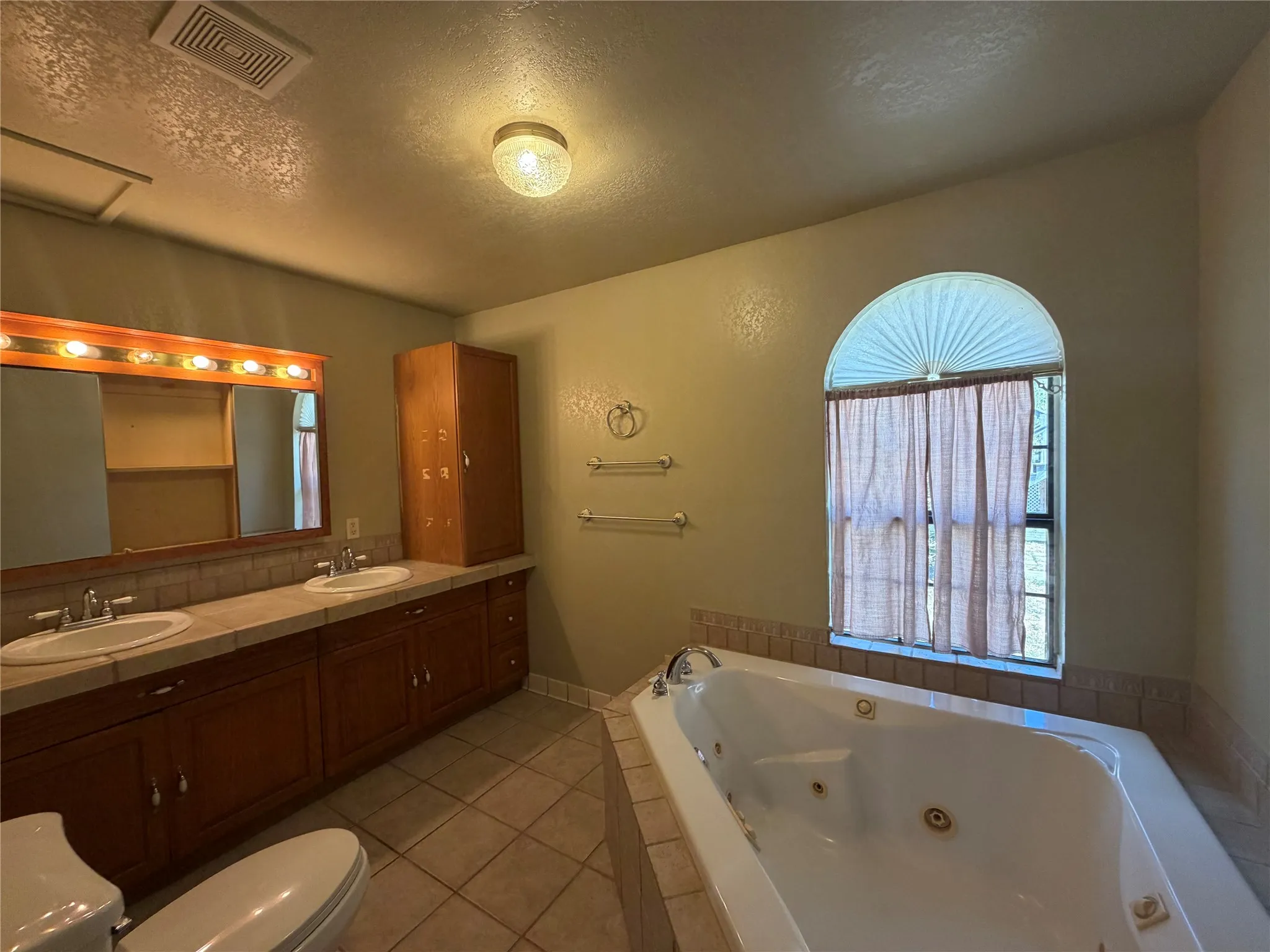 Dual vanity with integrated sinks, tiled countertop, wood-finish cabinetry, jetted soaking tub, and arched window
