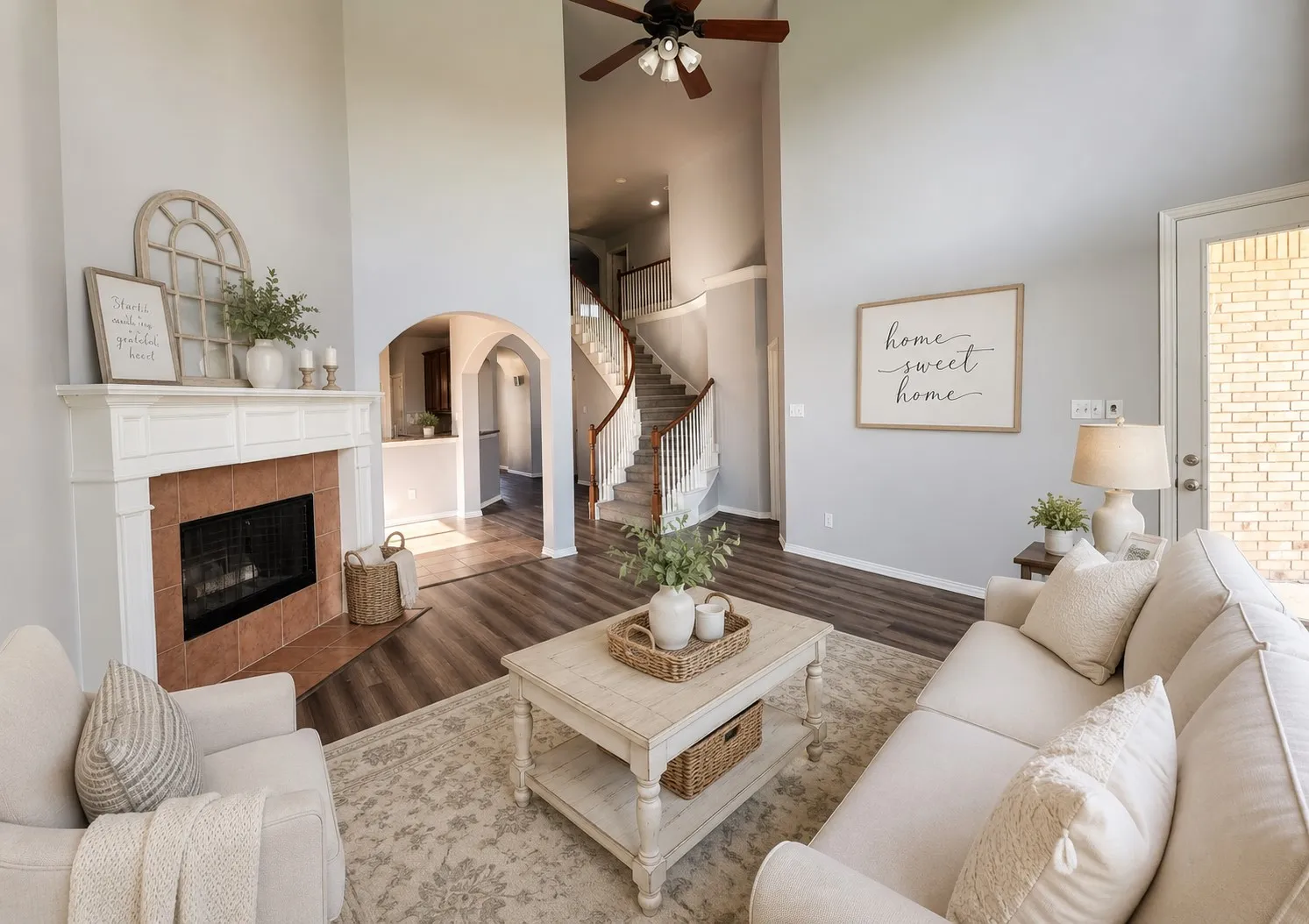 Two-story living area with high ceilings, a tiled fireplace with white mantel, plank flooring, a ceiling fan, and a light-colored exterior door