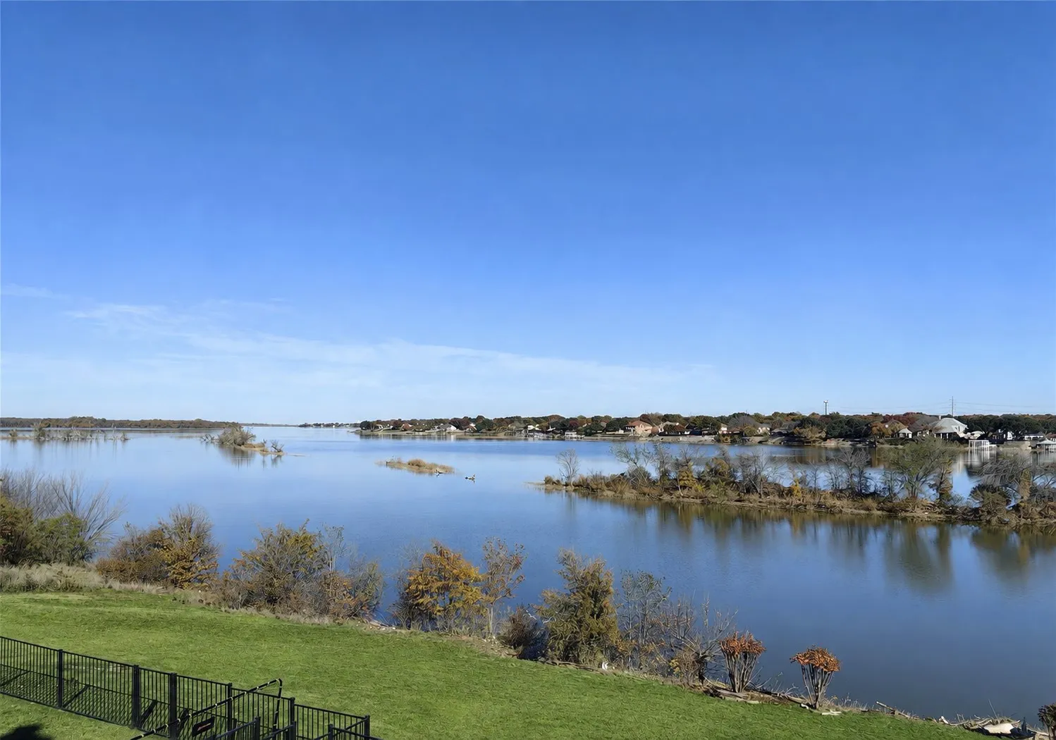 Expansive waterfront property with a manicured lawn, secure perimeter fencing, and mature landscaping