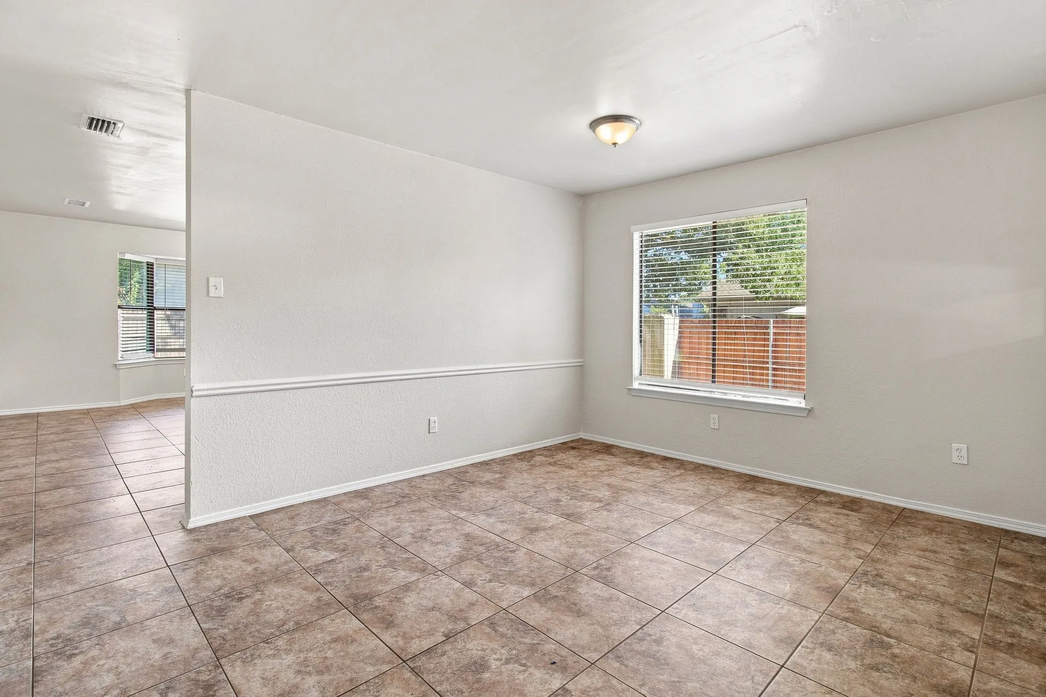 Spacious room featuring neutral tile flooring, a large window with blinds, and ceiling-mounted lighting