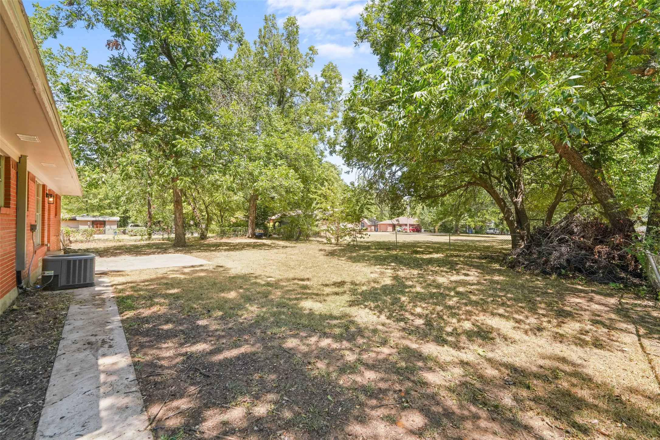 Expansive backyard with mature trees providing ample shade, a concrete patio, and a concrete pathway leading alongside the brick exterior