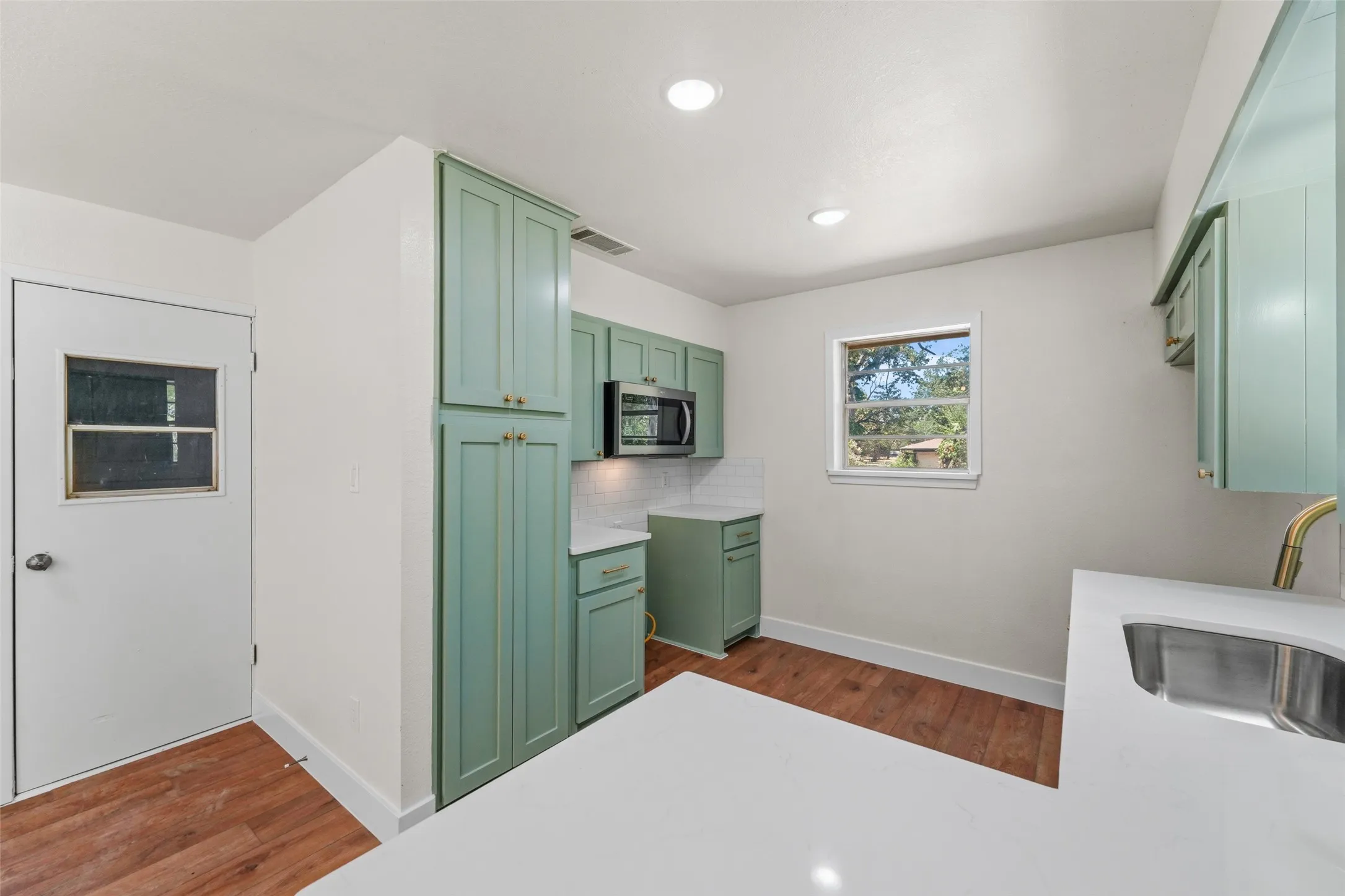 Kitchen featuring light green cabinetry, stainless steel microwave, white countertops, a subway tile backsplash, and wood-finish flooring