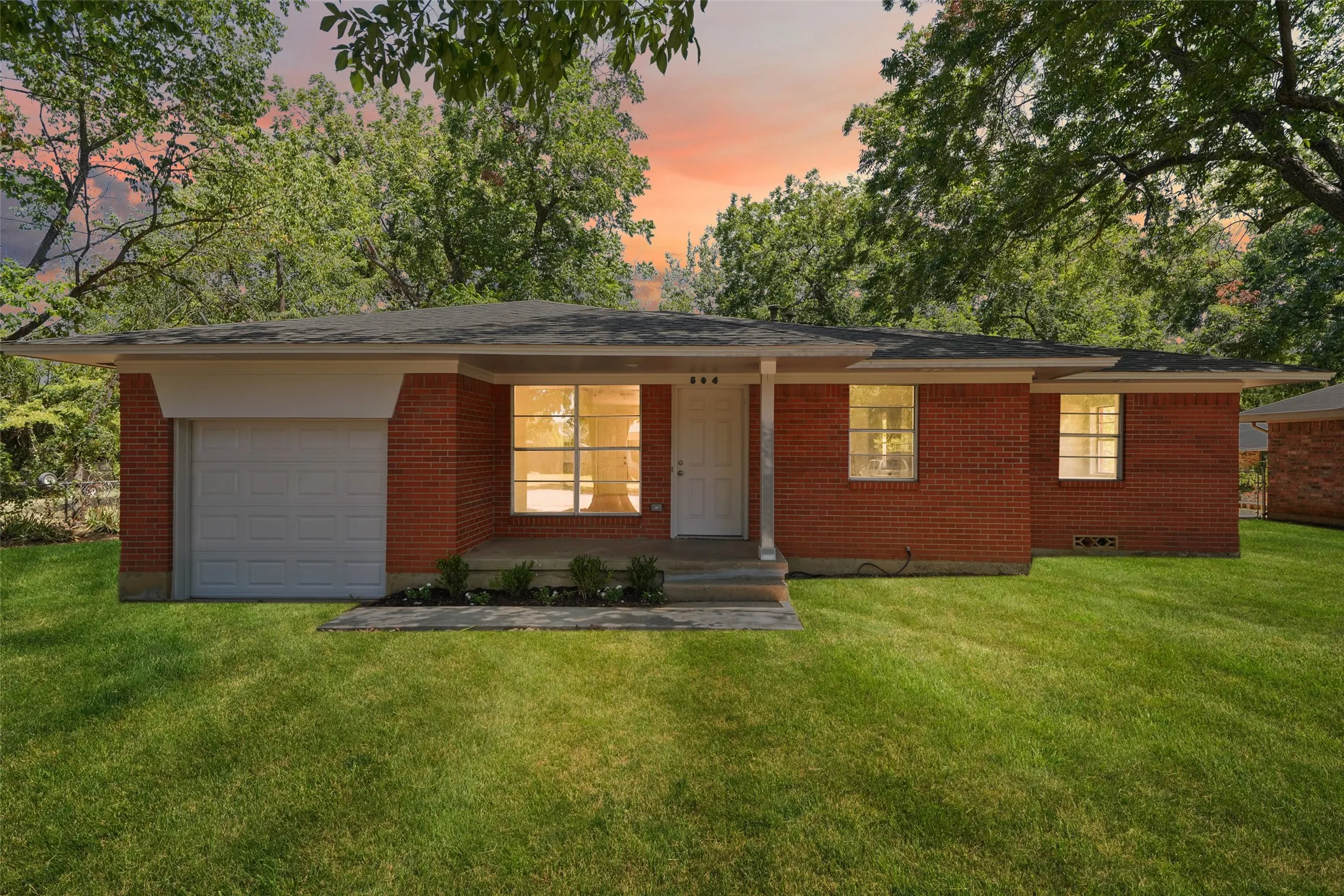 Brick exterior with a single-car garage, covered porch entry, mature surrounding trees, and a manicured lawn