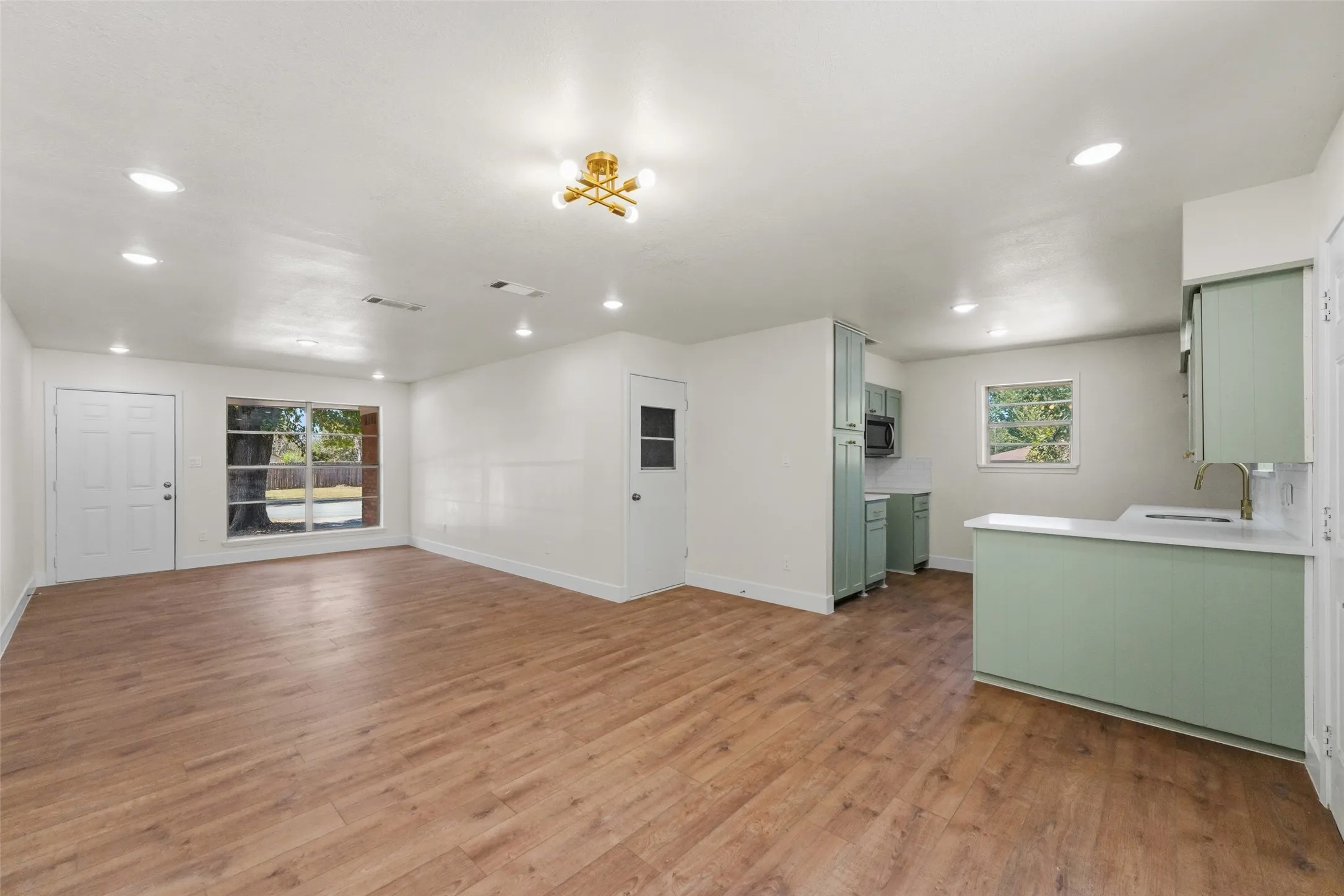 Open-concept living space featuring wood-finish flooring, recessed lighting, and a modern kitchen with pale green cabinetry, white countertops, and a built-in microwave