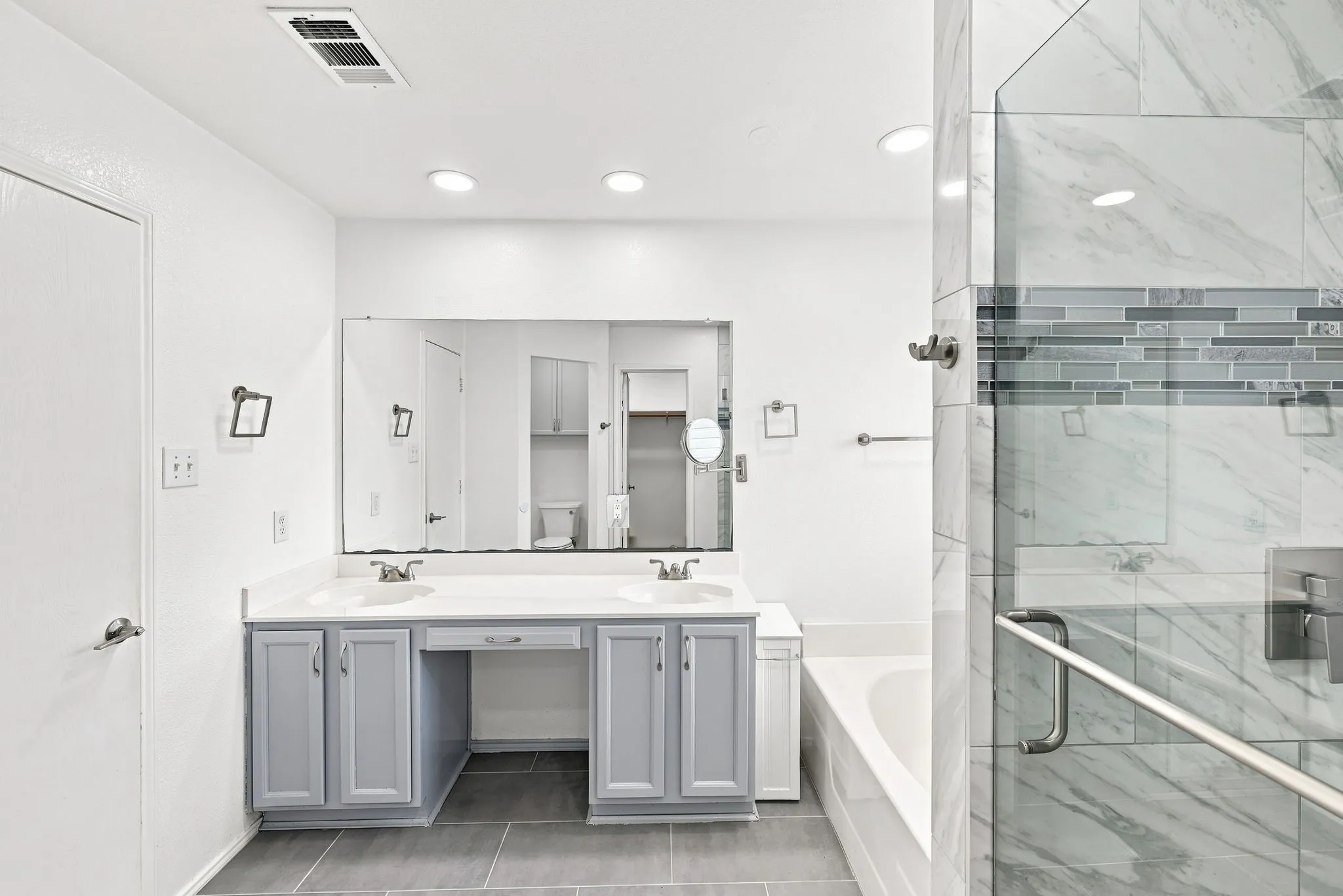 Dual vanity with gray cabinetry and white countertops, large wall-mounted mirror, recessed lighting, glass-enclosed shower with tiled surround, and separate soaking tub