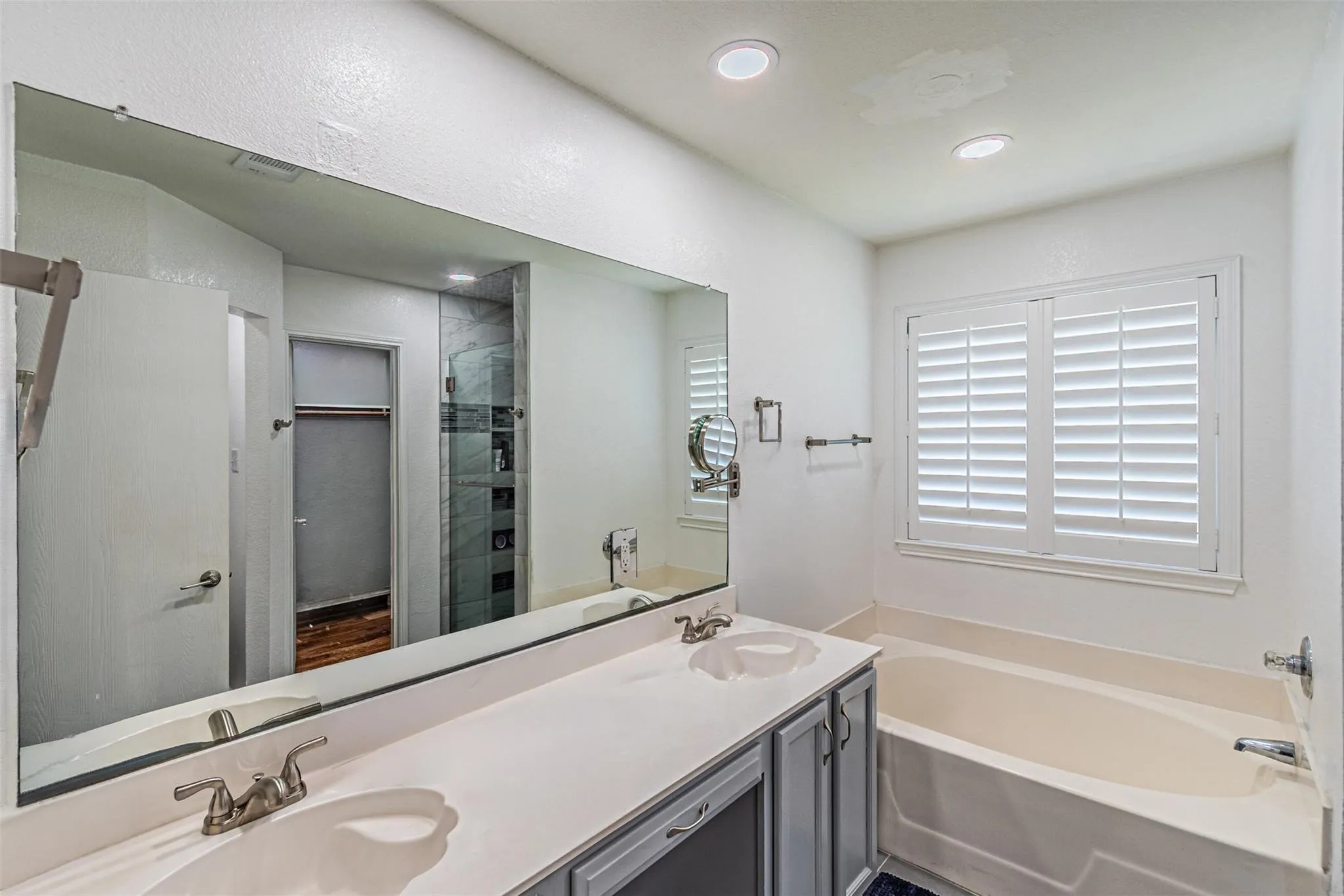 Dual vanity with integrated sinks, full-width mirror, recessed lighting, soaking tub, and window with plantation shutters
