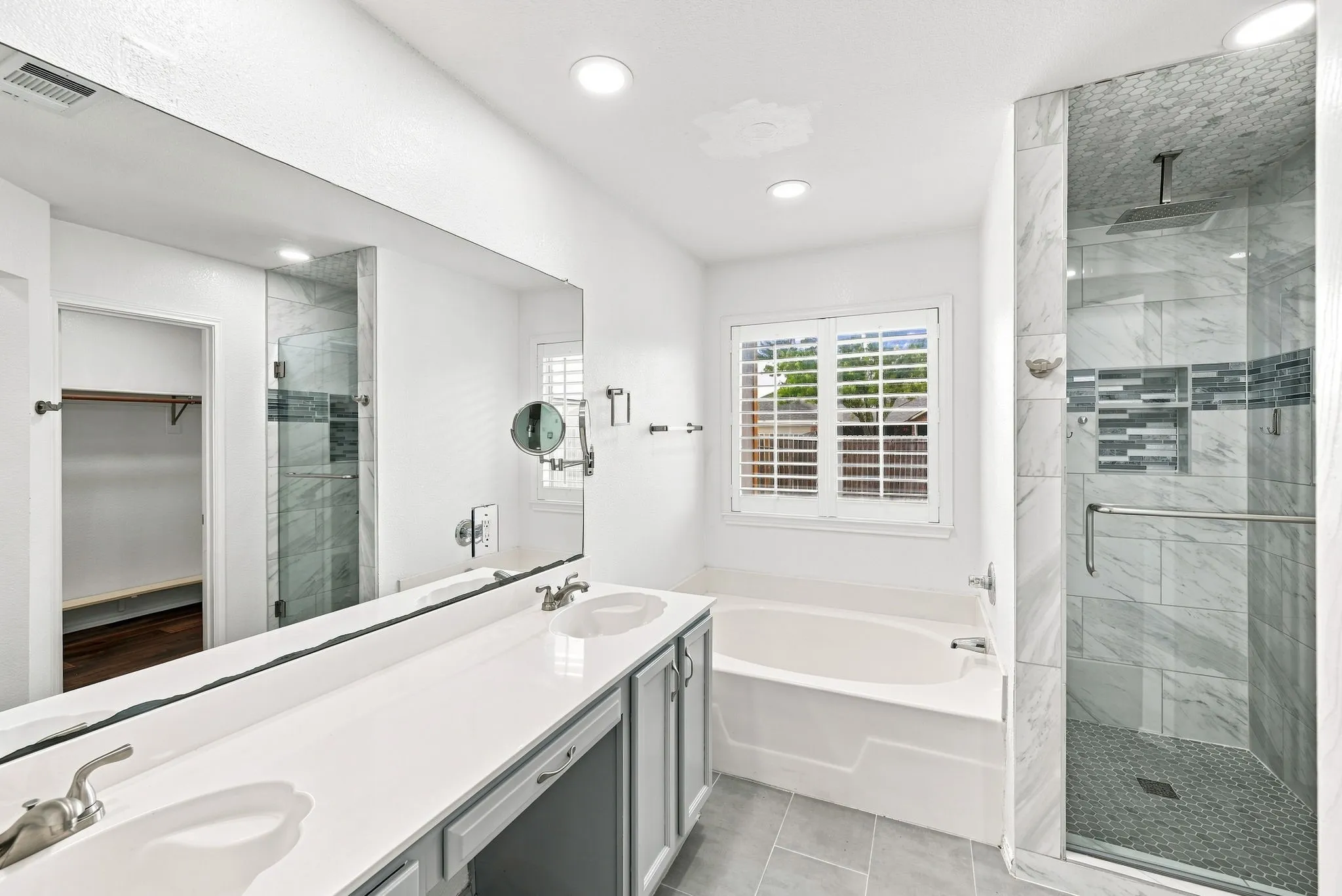 Dual vanity with undermount sinks, separate shower with glass enclosure and mosaic tile accents, built-in bathtub, and window with plantation shutters