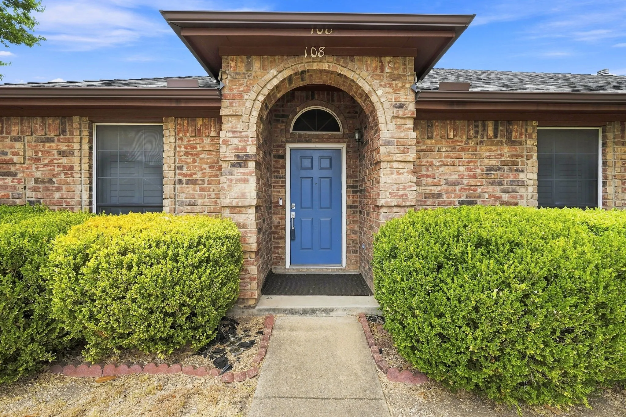 Brick exterior with an arched entryway, featuring a blue paneled front door, an asphalt shingle roof, and mature perimeter landscaping