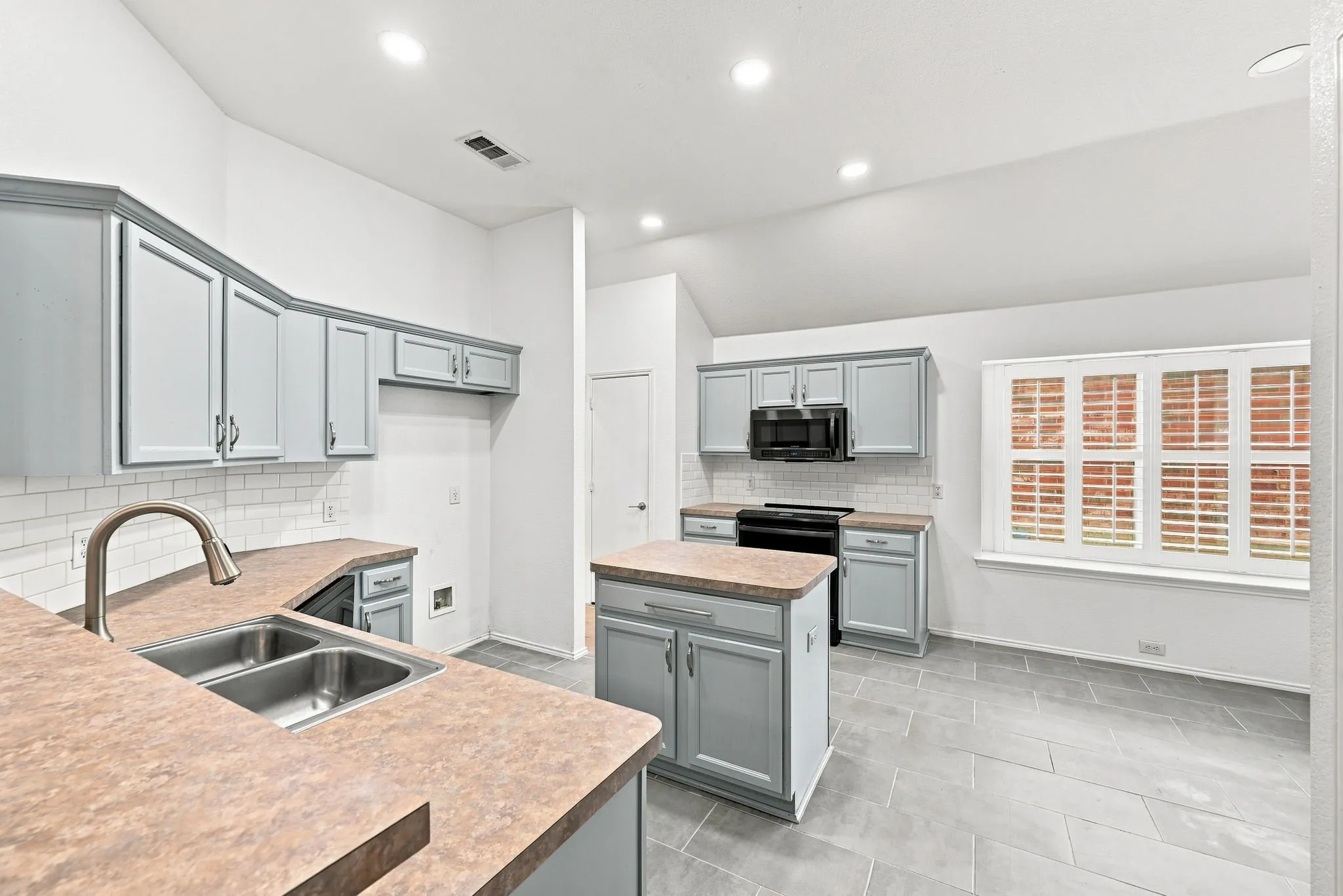 Kitchen featuring a dual-basin sink, light-toned cabinetry, a central island, tiled flooring, and a window with plantation shutters
