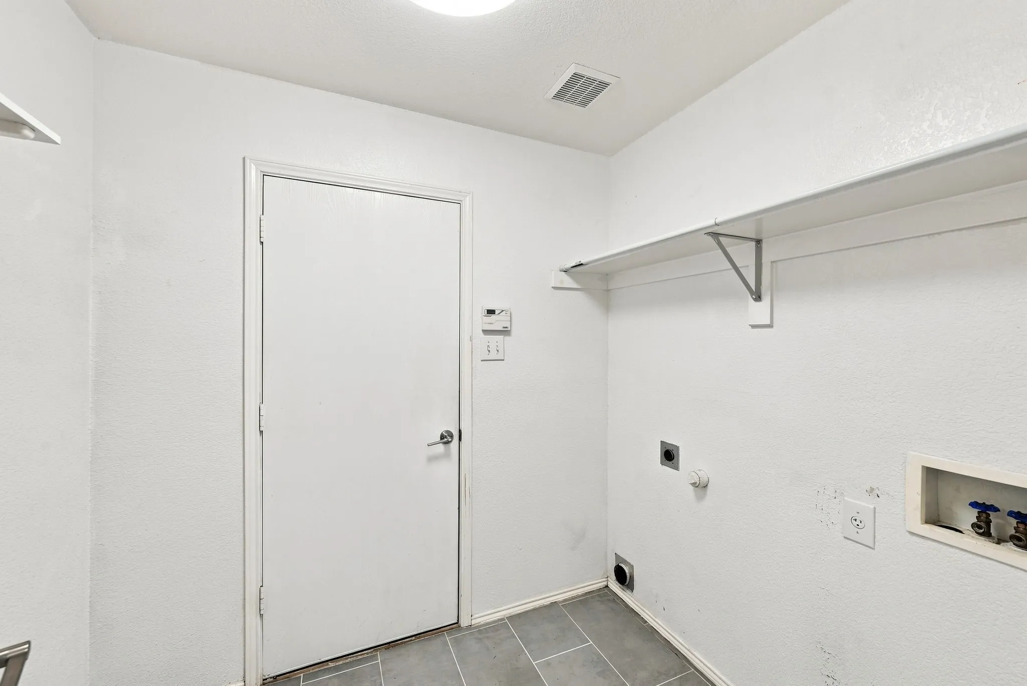 White walls with an overhead shelf, utility connections, a closed door, and gray tile flooring