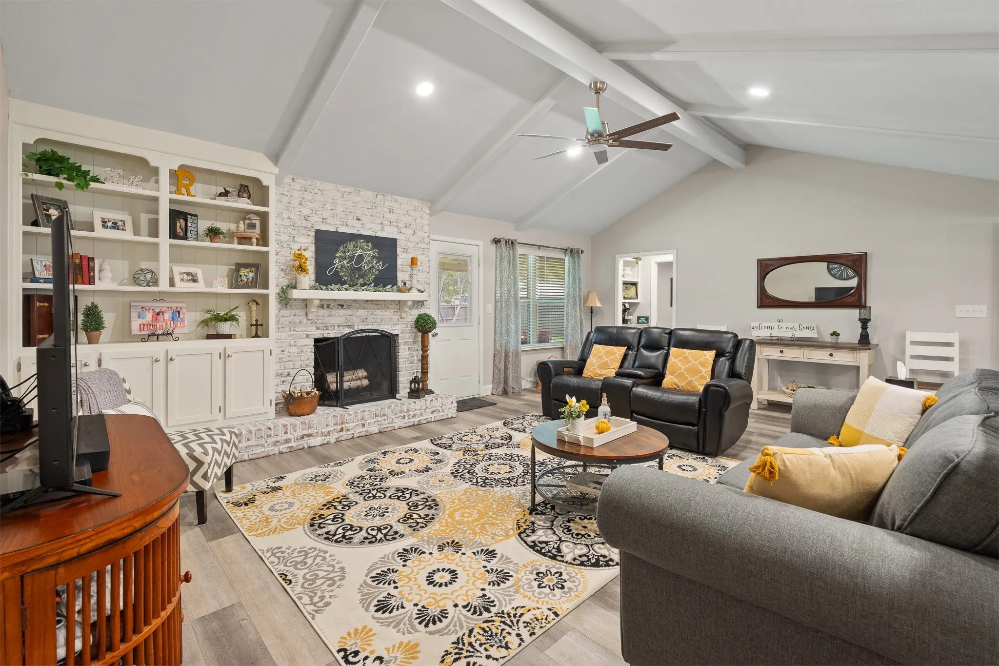 Vaulted ceiling with exposed beams, wood-finish flooring, built-in shelving, brick fireplace with mantel, and a ceiling fan