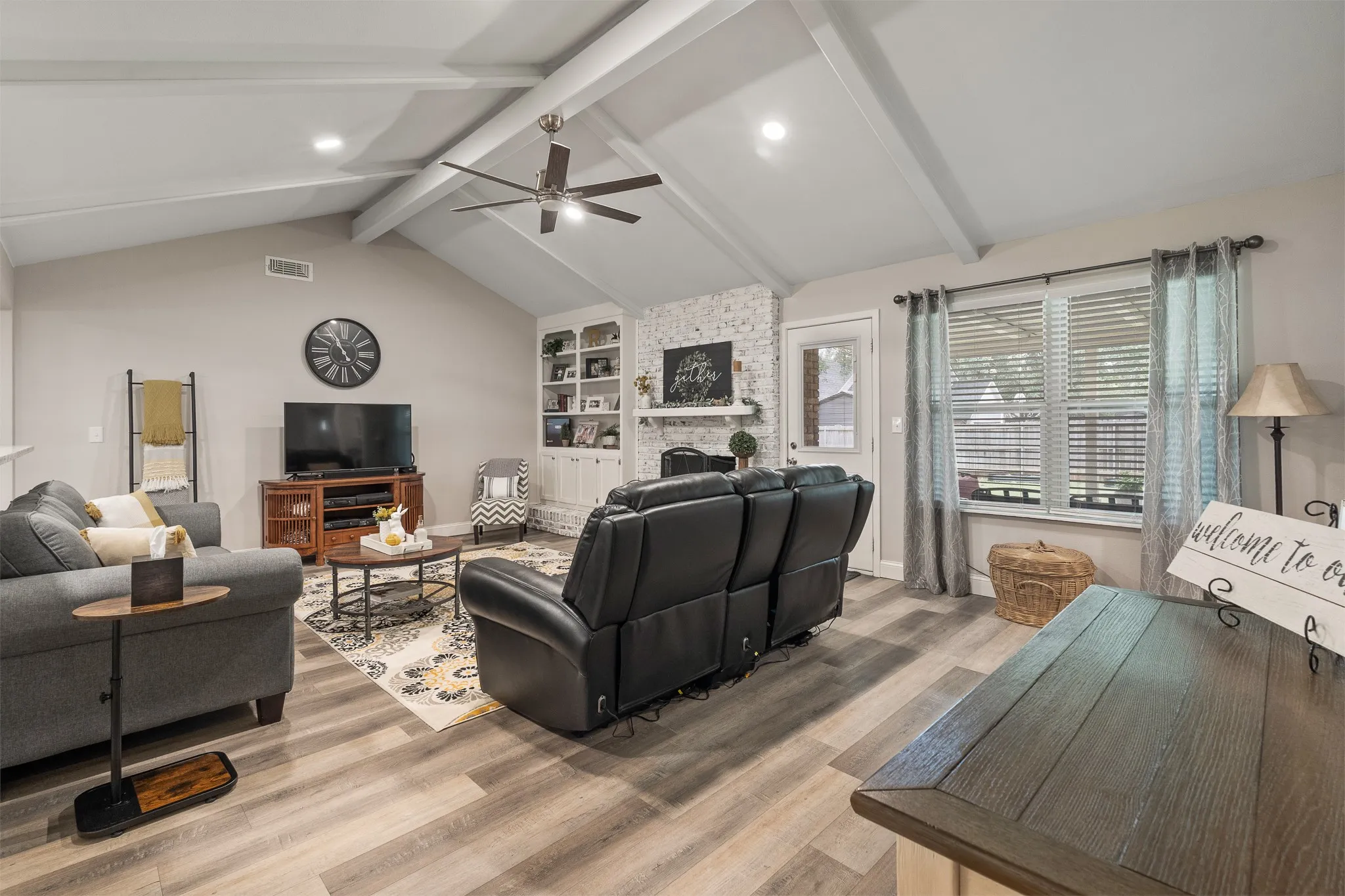 Vaulted ceiling with exposed beams, wood-finish flooring, built-in shelving, stone-finish fireplace, and ceiling fan