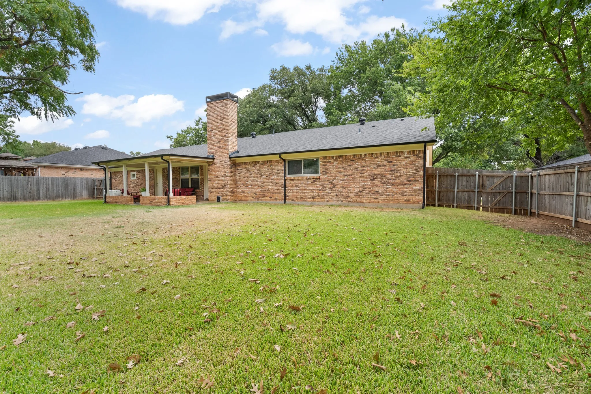 Brick exterior with a covered patio, a prominent chimney, a dark shingled roof, and a wood privacy fence