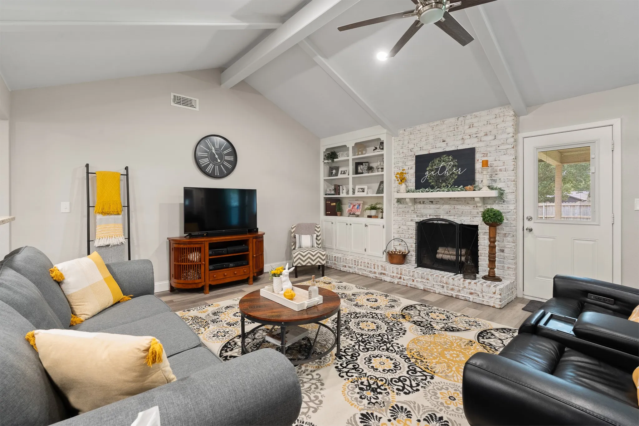 Vaulted ceiling with exposed beams, wood-finish flooring, white brick fireplace, built-in shelving, and a ceiling fan