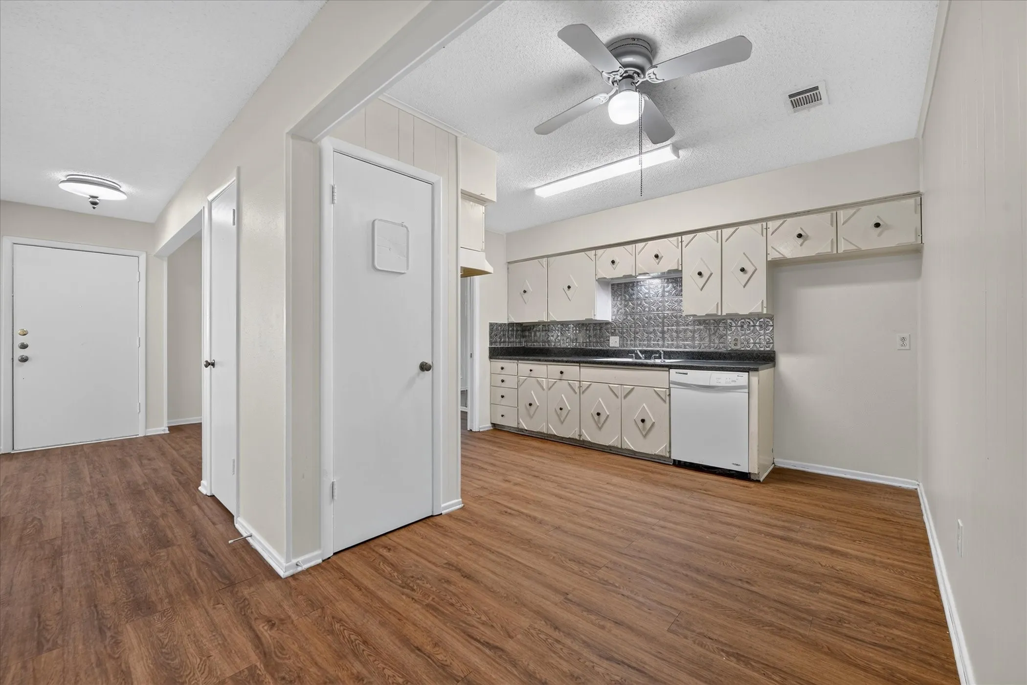 Wood-finish flooring throughout, white paneled doors, and a kitchen featuring white cabinetry with dark hardware, a dark countertop, and a patterned tile backsplash