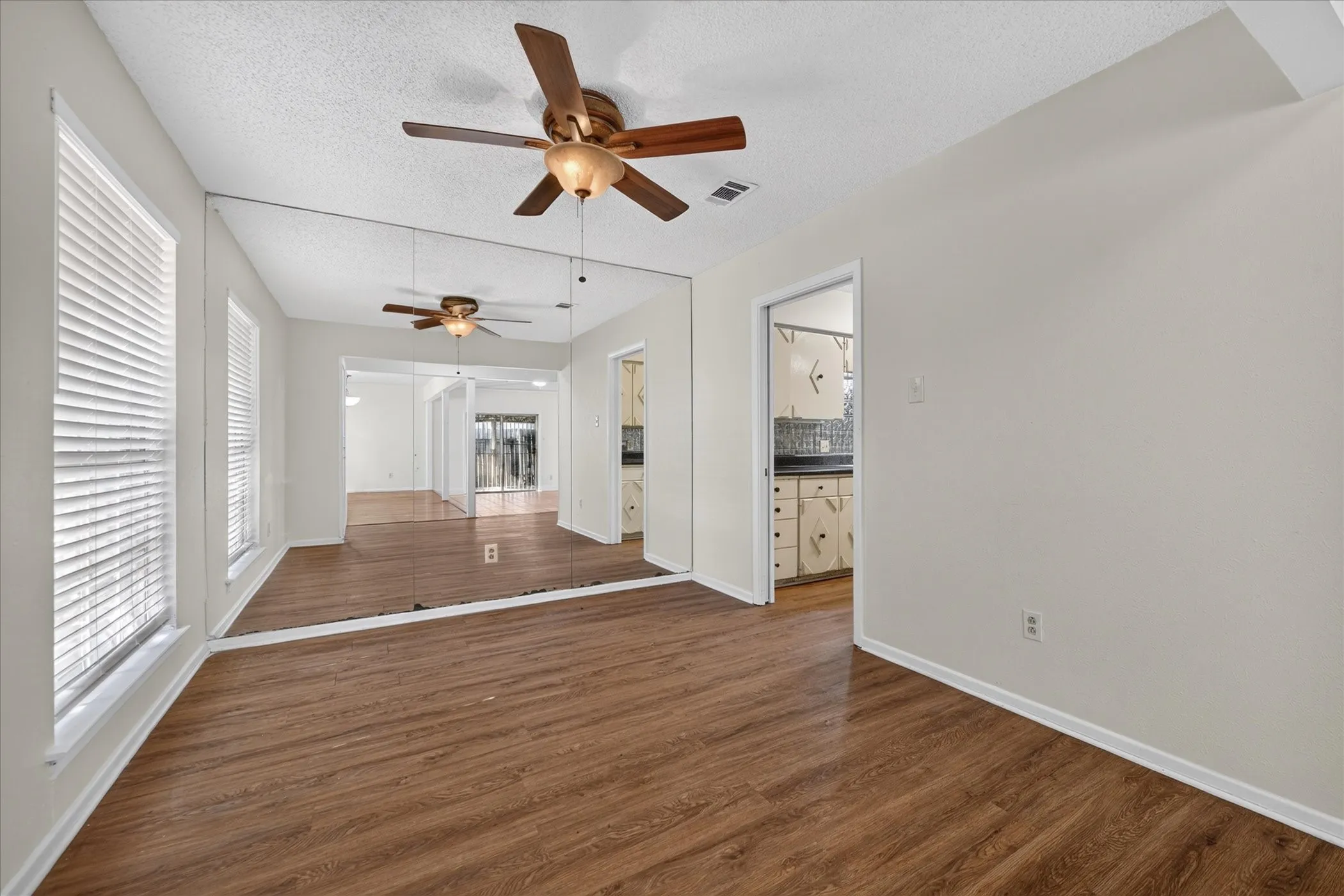 Wood-finish flooring, two windows with blinds, a ceiling fan with integrated lighting, and a mirrored accent wall