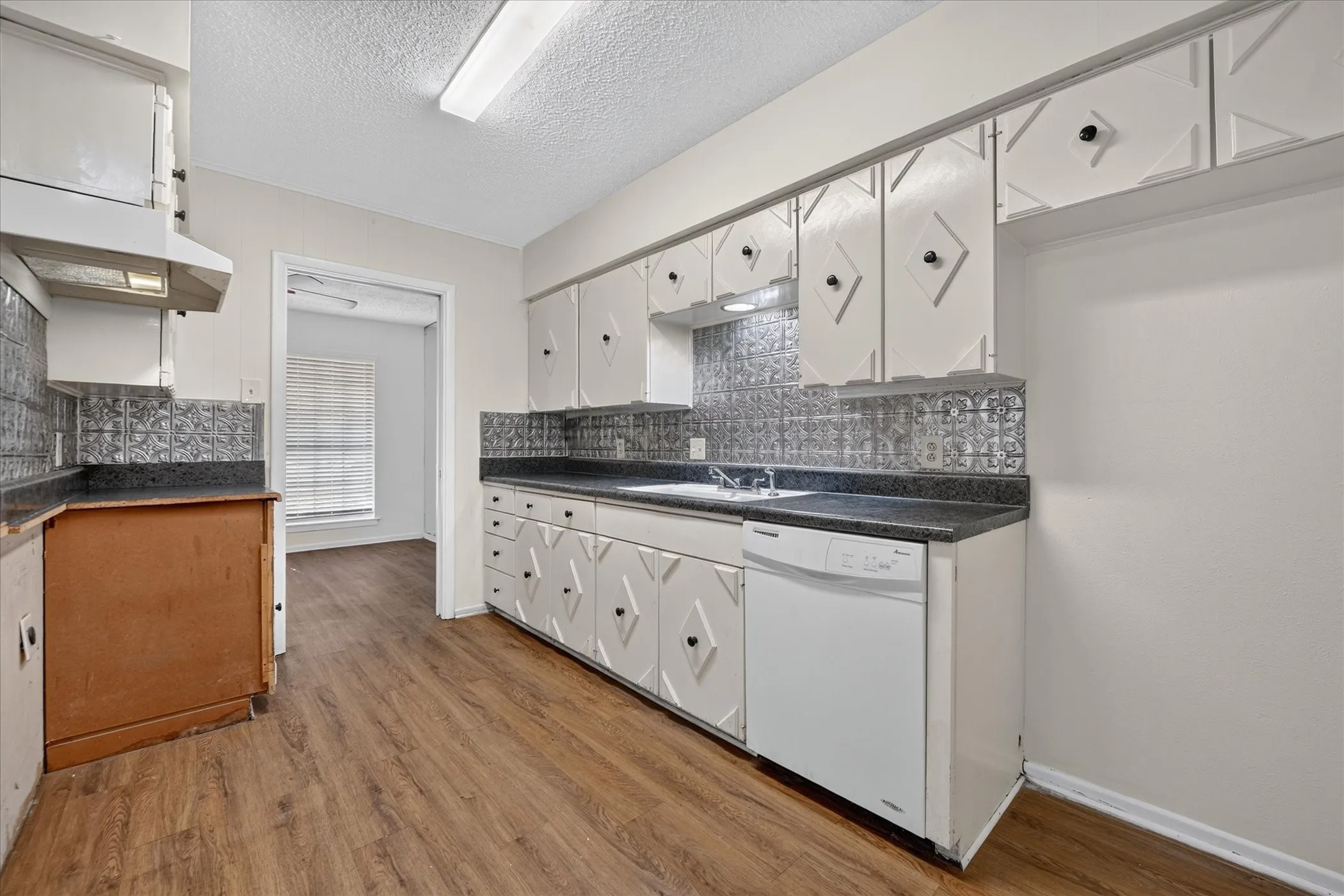 Kitchen featuring wood-finish flooring, white cabinetry with decorative panels, dark countertops, a patterned tile backsplash, and a white dishwasher