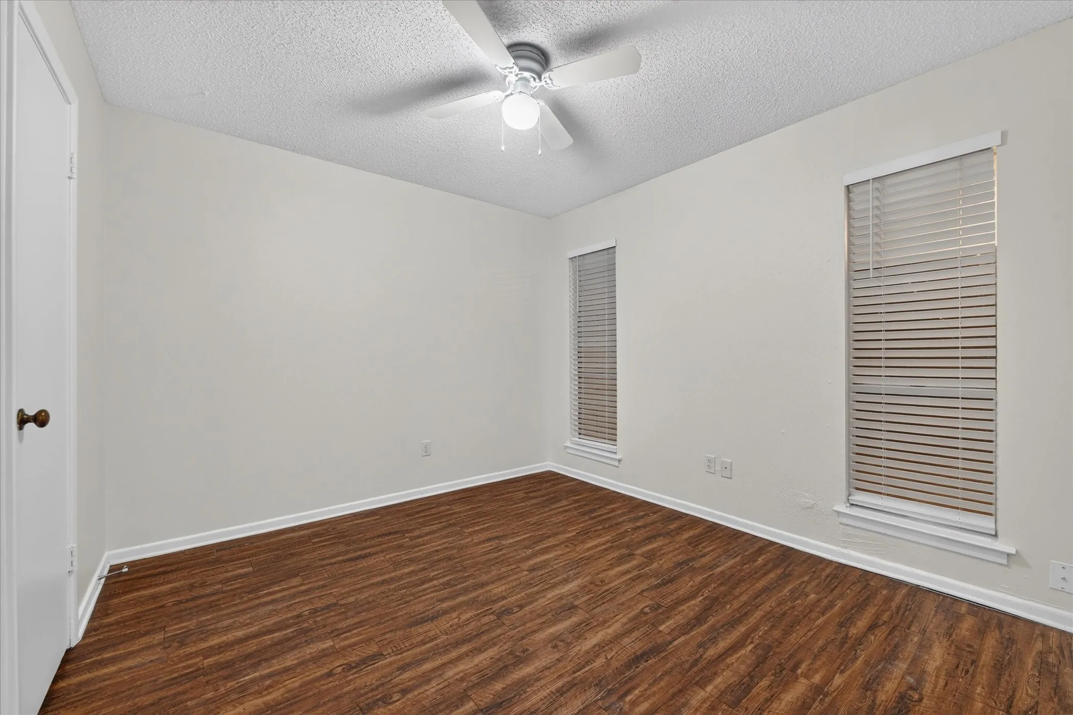 Wood-finish flooring with white baseboards, two windows with blinds, a ceiling fan, and a white door with a dark knob