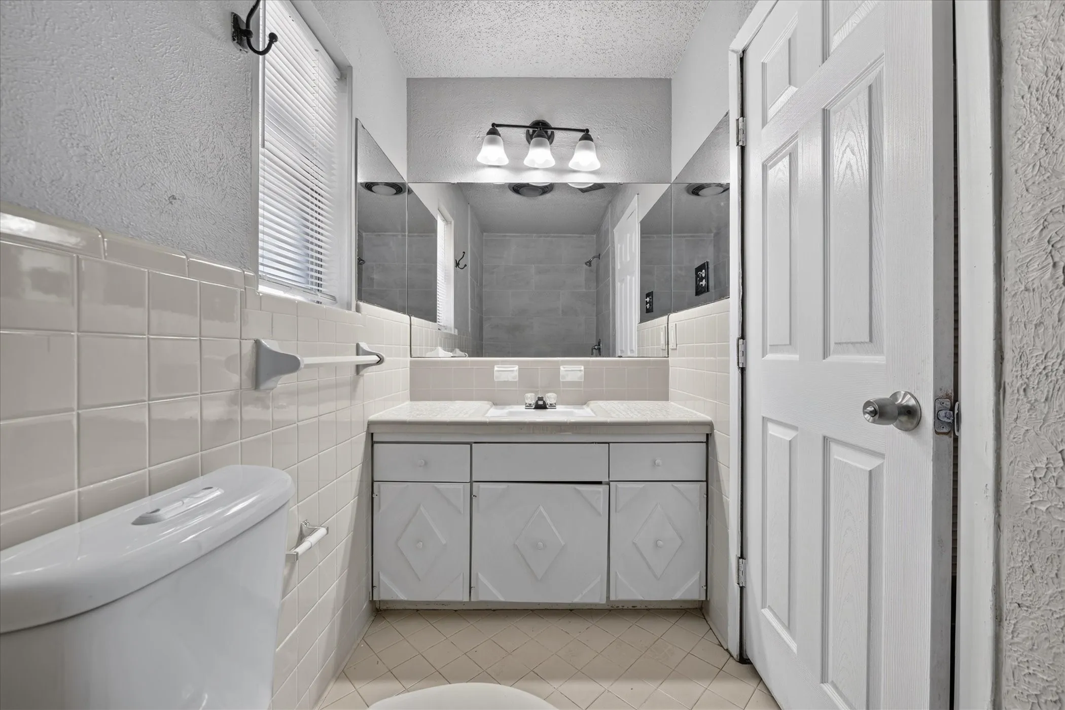 Bathroom featuring a white vanity with integrated sink, wall-mounted mirror, three-light fixture, and tiled walls