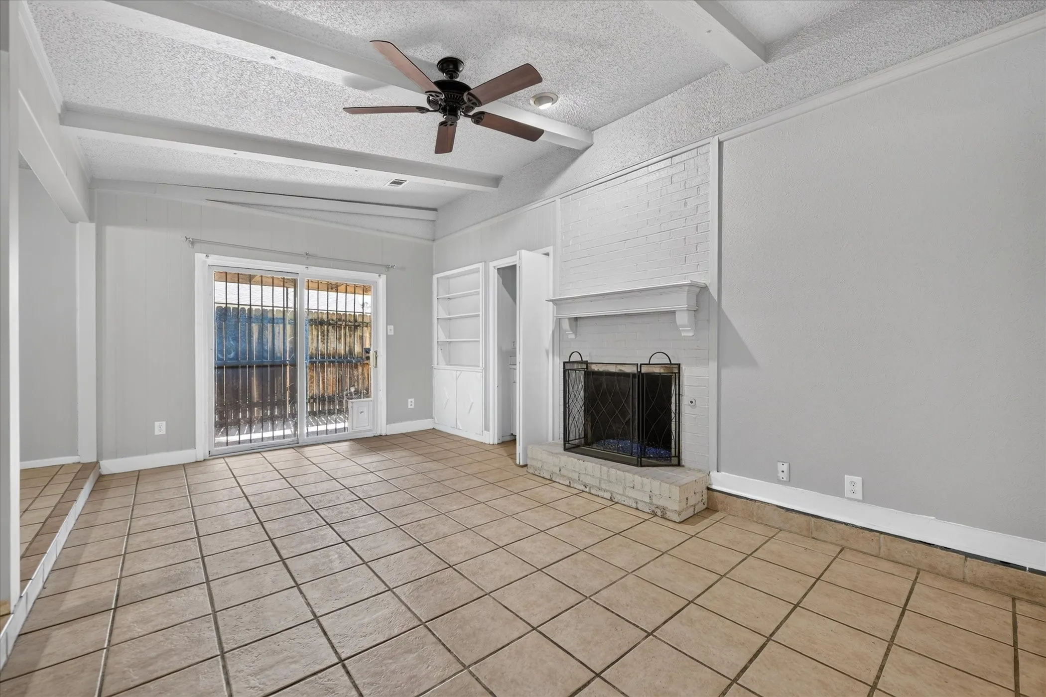 Spacious room featuring tile flooring, exposed beam ceiling, a ceiling fan, a brick fireplace with a mantel, and built-in shelving
