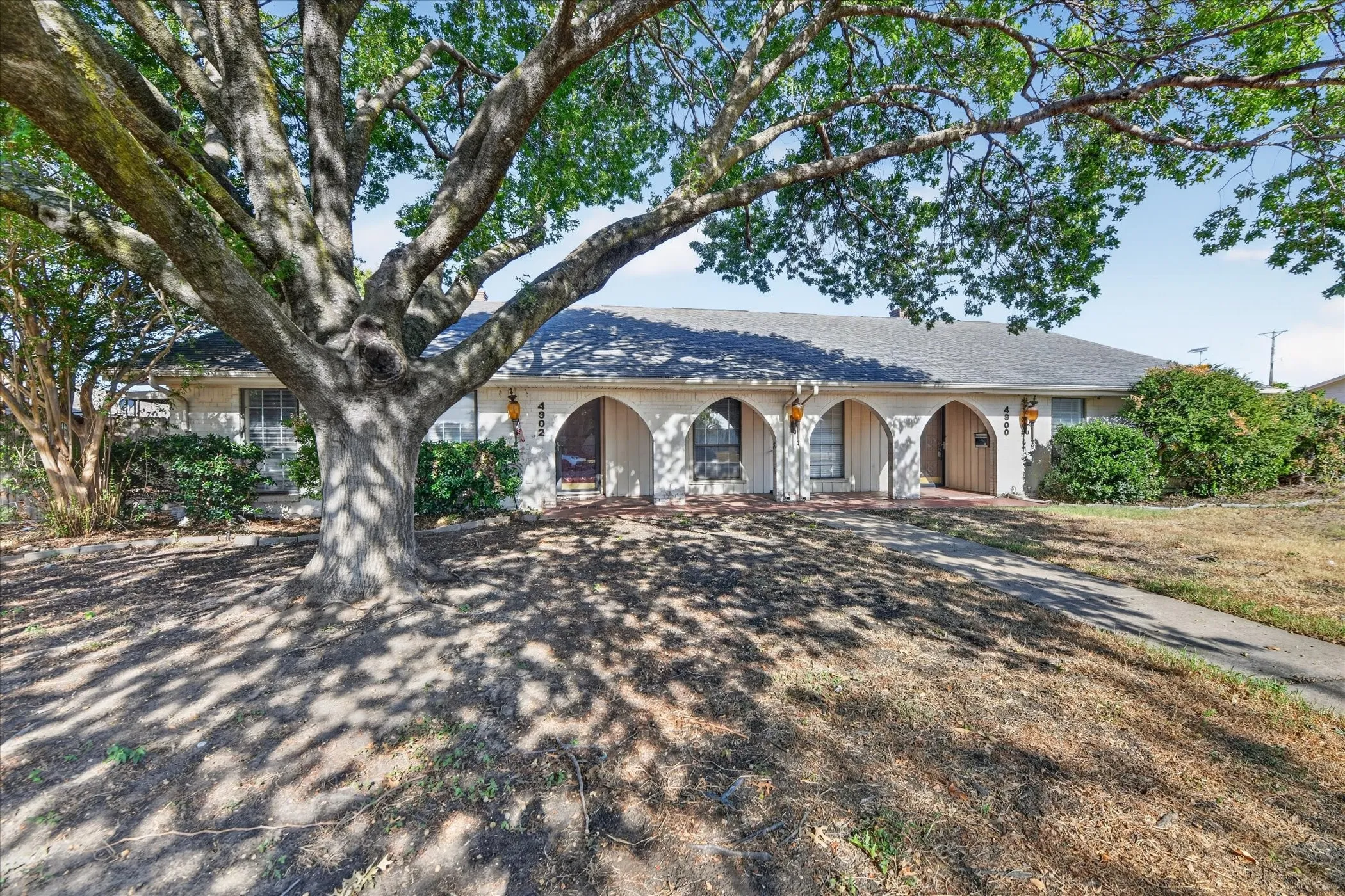 Single-story structure with a gray shingle roof, featuring multiple arched entryways and a mature tree with extensive canopy